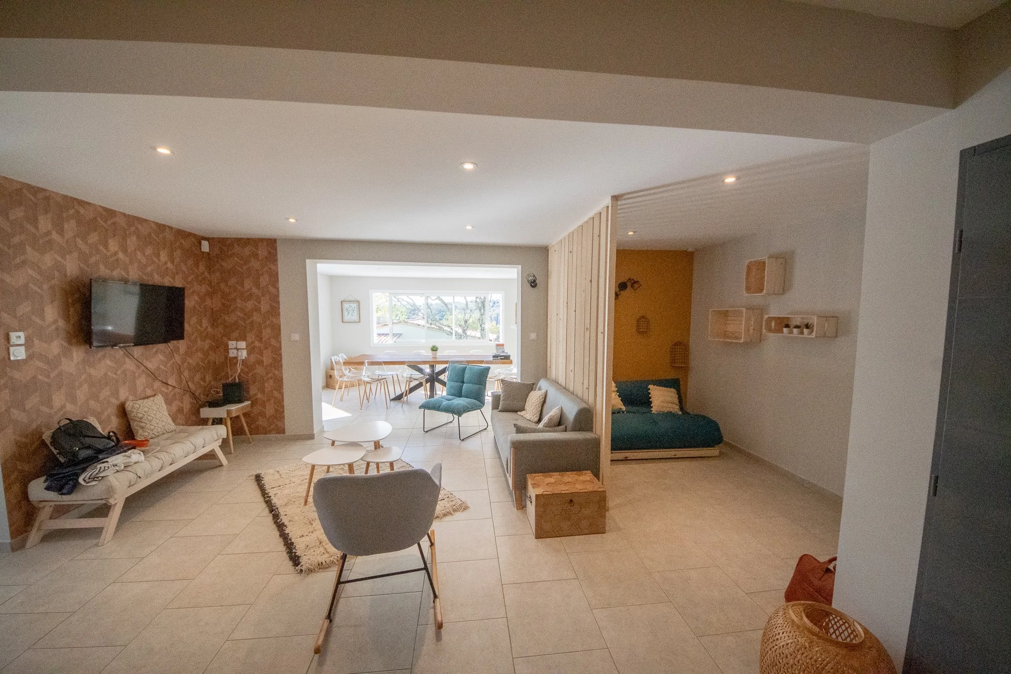 Salon avec canapé gris, fauteuil bleu, télévision murale, table basse, mur en bois, zone de lecture avec un canapé vert, buffet blanc, espace lumineux avec grande fenêtre, décoration minimaliste