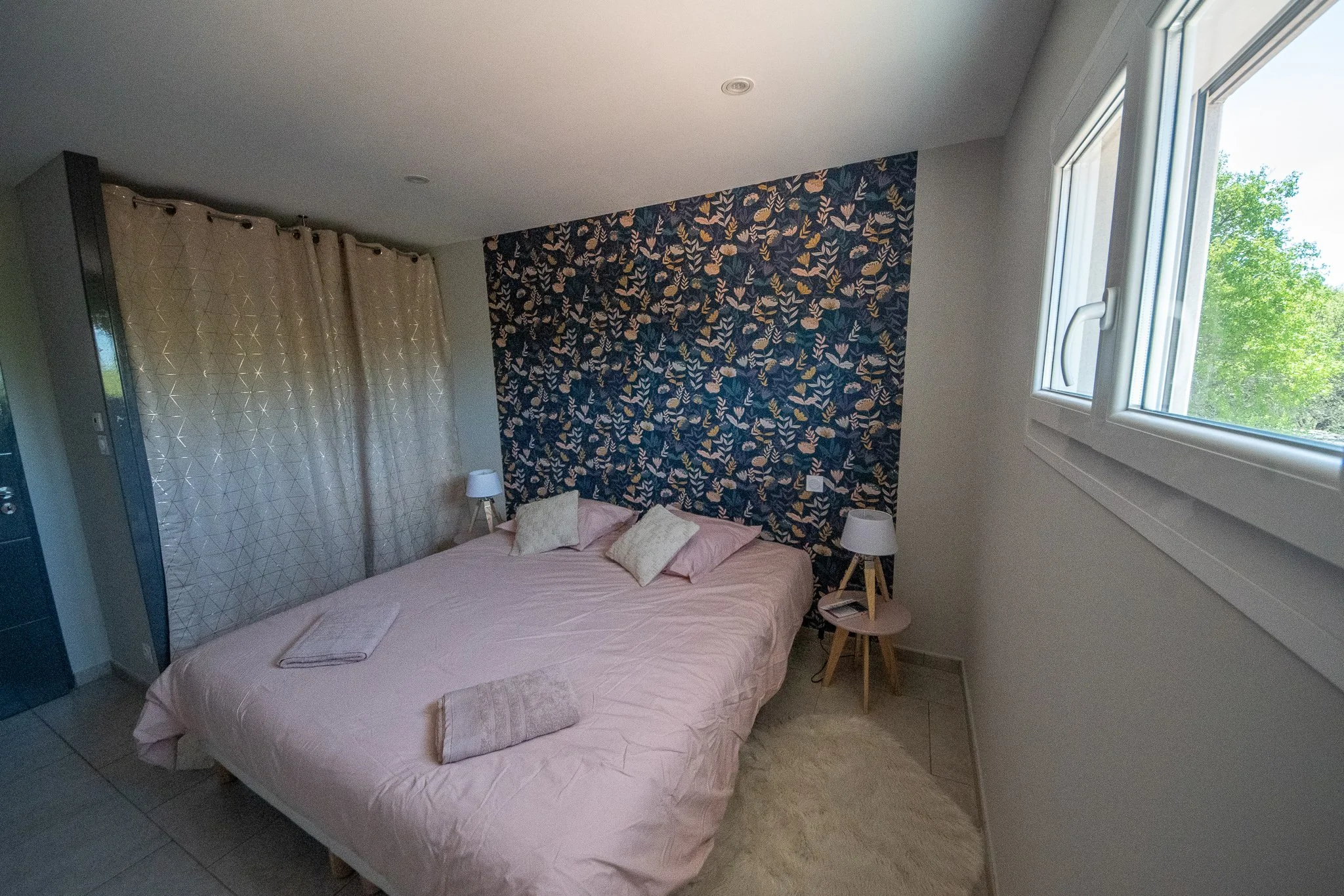 Chambre avec lit double, coussins, lampes de bedside, mur avec papier peint à motif floral, fenêtre laissant entrer de la lumière naturelle.