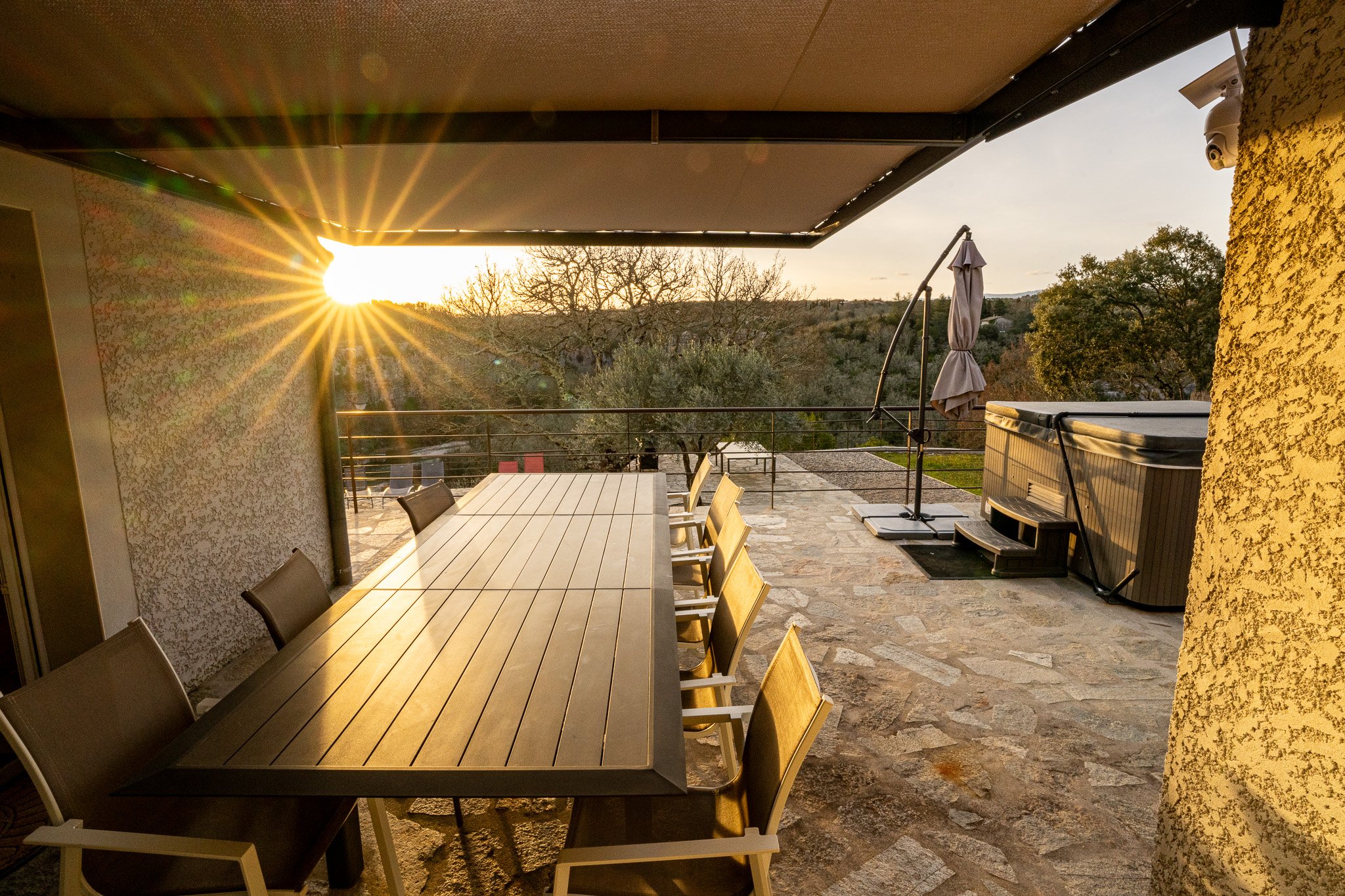 Pergola abritant la terrasse avec table et chaises sous un manteau de soleil couchant, vue sur des arbres et un jacuzzi.