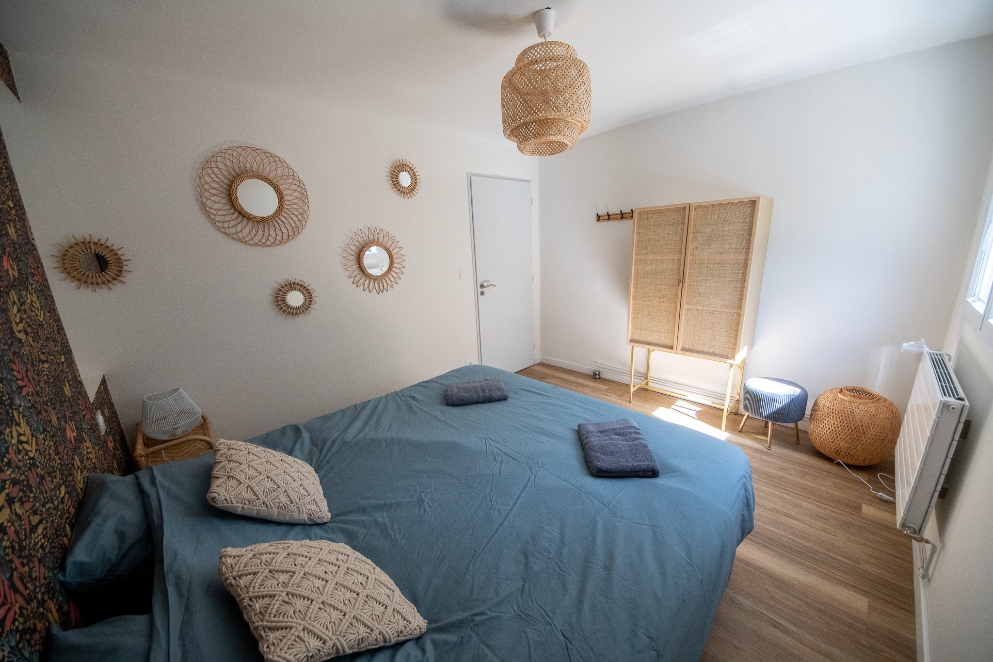 Chambre avec un lit double, drap bleu, deux serviettes pliées, oreillers en coussin texturé, décoration en rotin, miroir, ambiance lumineuse naturelle.