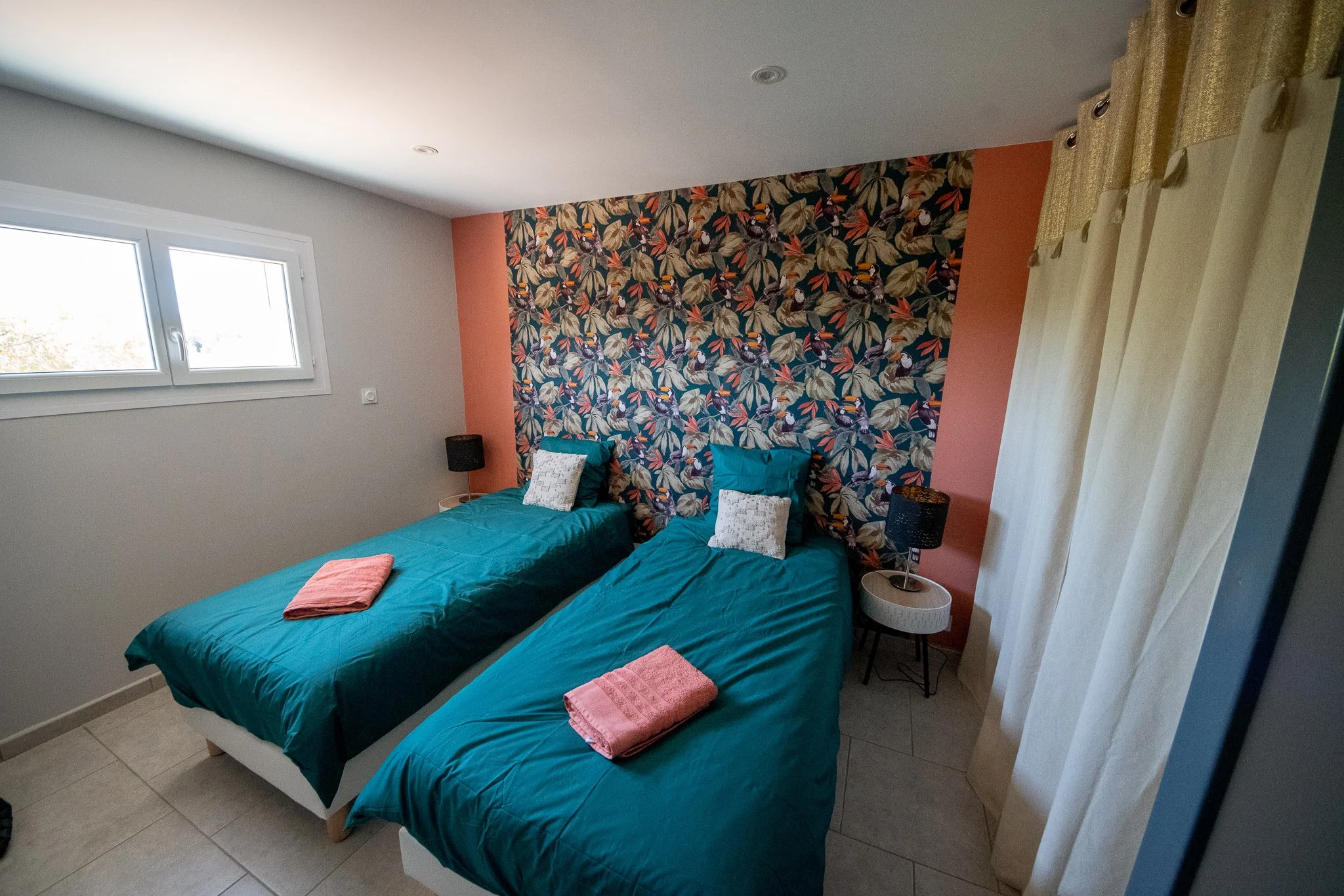 Chambre avec deux lits, une tapisserie florale en arrière-plan, deux lampes de nuit, deux têtes de lit bleues, deux serviettes de bain roses, une fenêtre avec rideaux blancs.