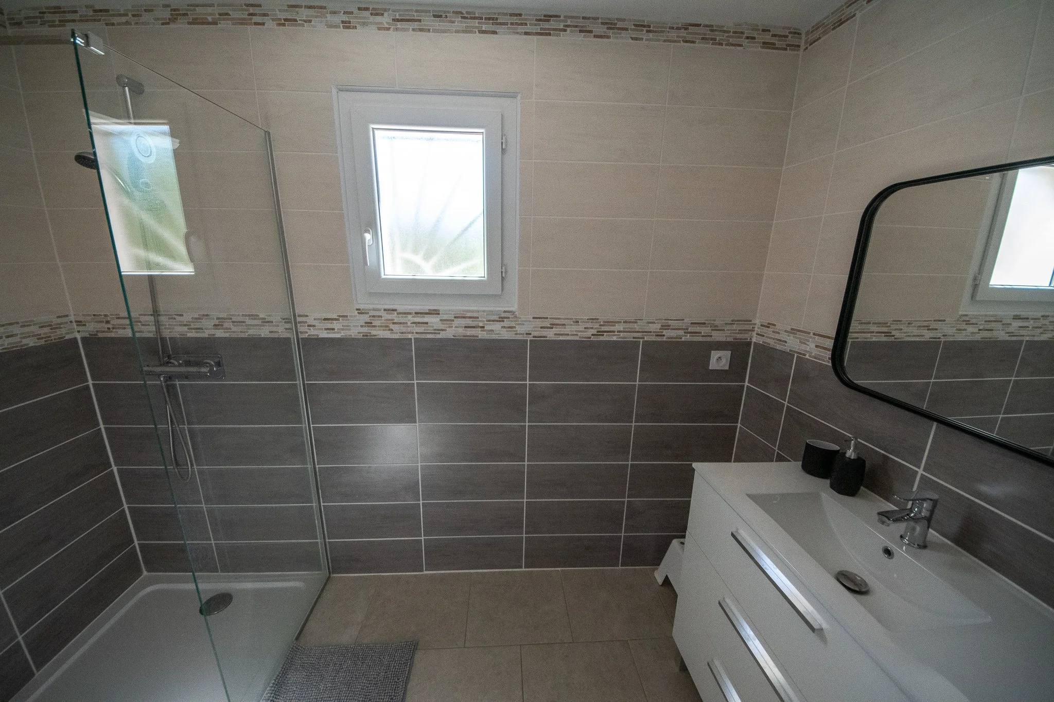 Salle de bain moderne avec douche à l'italienne, petite fenêtre, miroir rectangulaire, meuble de lavabo blanc avec deux tiroirs, meuble noir avec savon et brosse à dents, murs carrelés gris et beige, tapis de bain gris