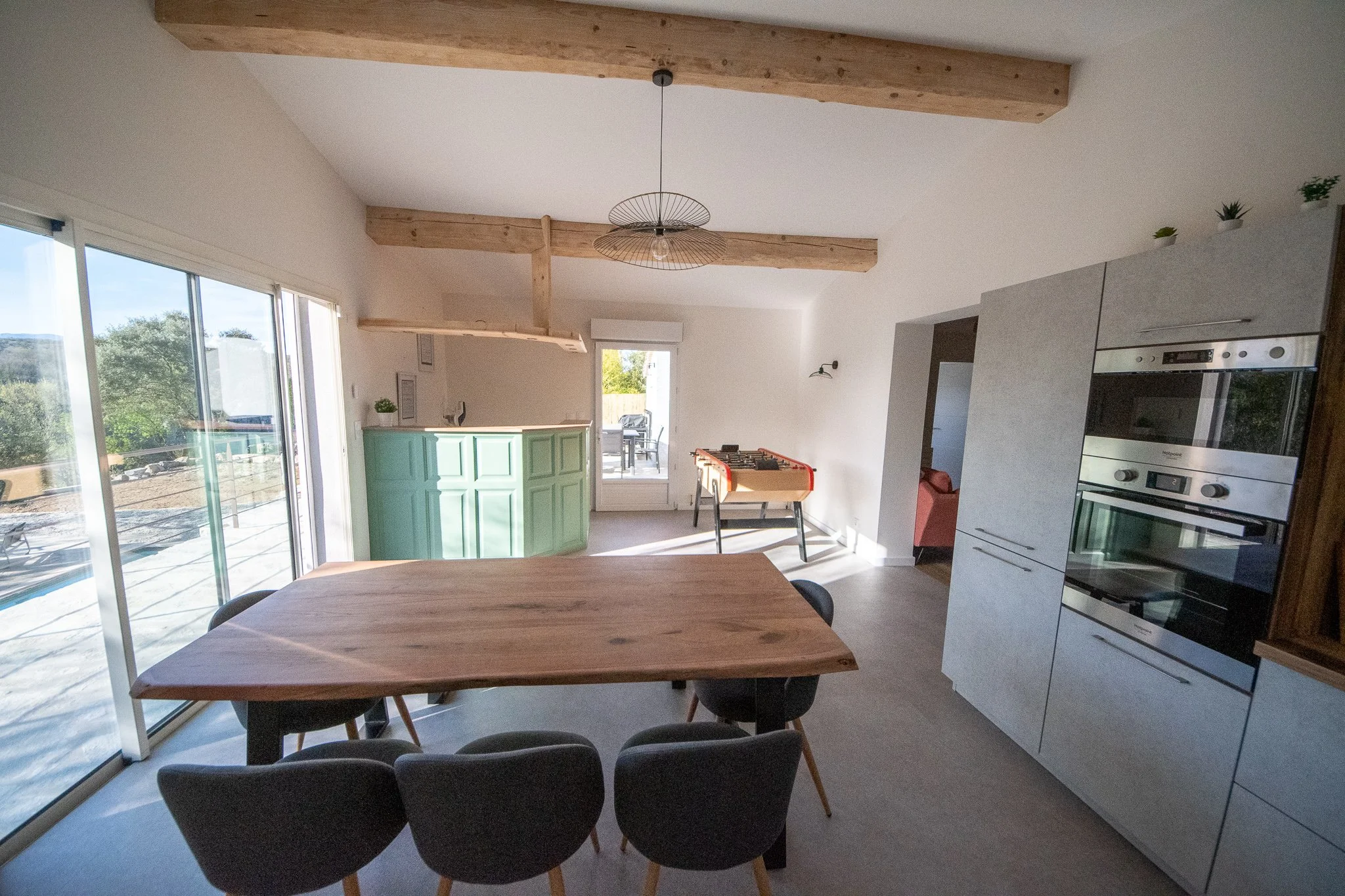 Intérieur d'une cuisine moderne avec un espace salle à manger, grande baie vitrée, meubles en blanc et vert, plafond avec poutres en bois, terrasse extérieure visible, et un terrain naturel avec arbres