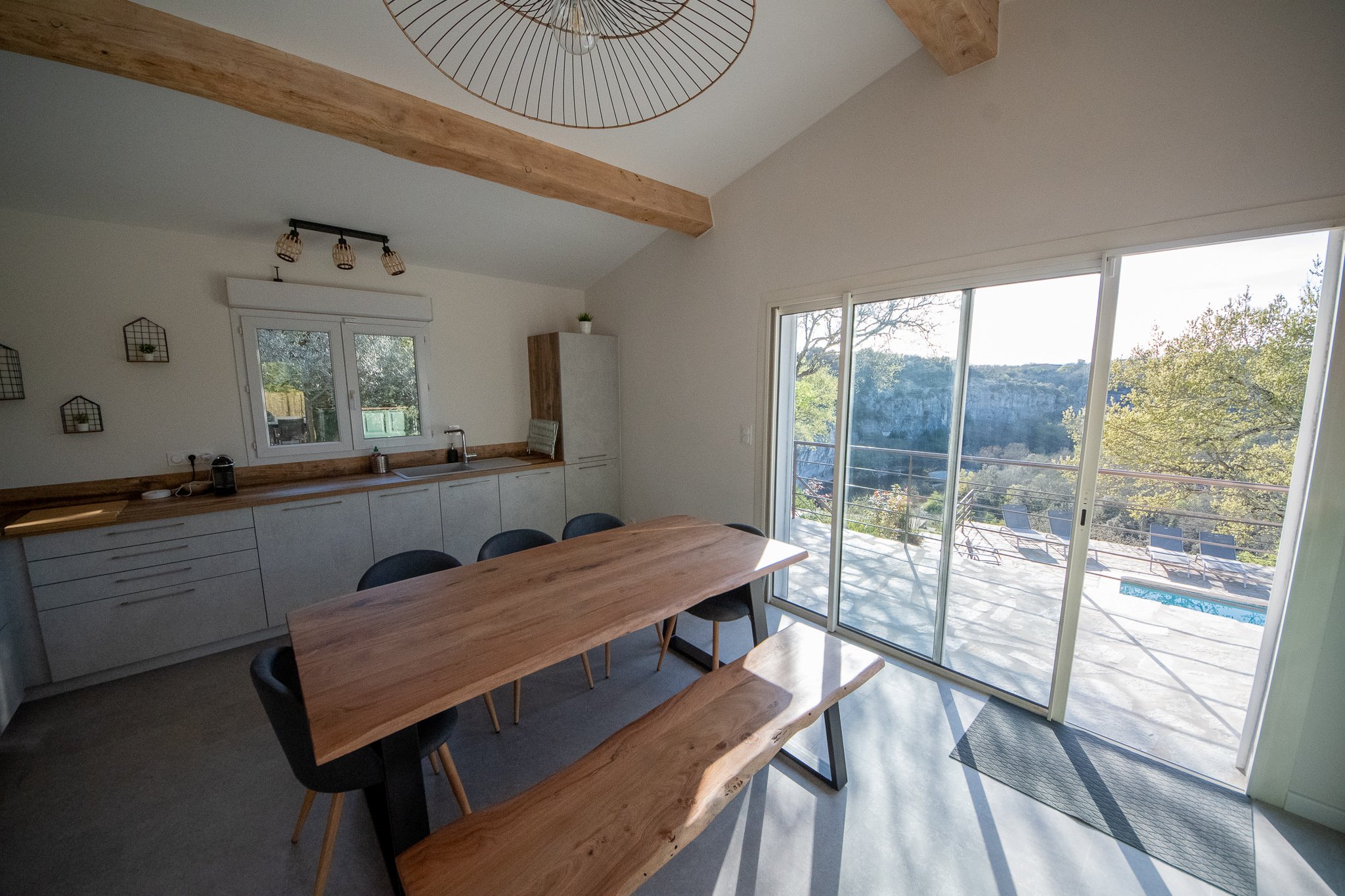 Salon de cuisine lumineux avec grande baie vitrée donnant sur une terrasse avec mobilier et piscine, meubles en bois clair, murs blancs, décoration minimaliste.