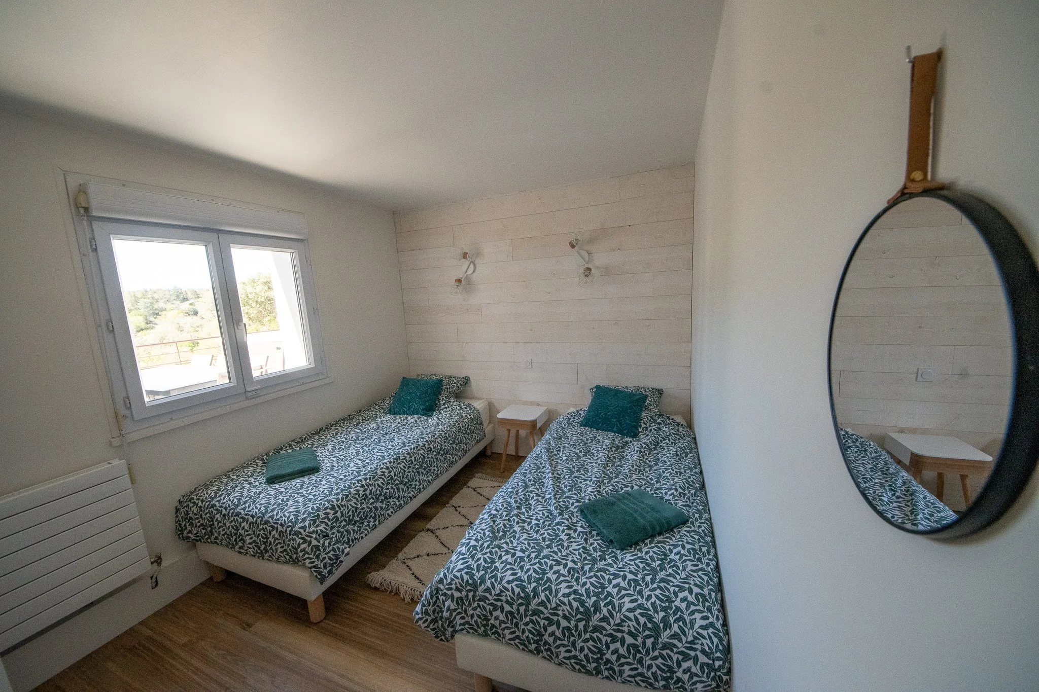 Chambre avec deux lits simples, deux coussins verts, tapis beige, mur en bois clair, miroir rond, fenêtre avec vue sur la nature.