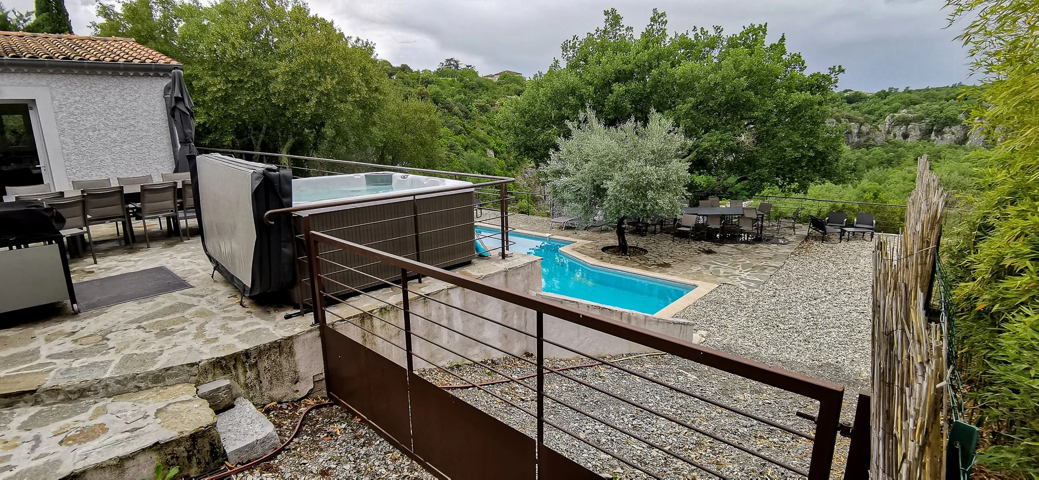 Vue d'une terrasse avec une piscine, un jacuzzi, plusieurs chaises et tables, entourée d'arbres et de collines verdoyantes.