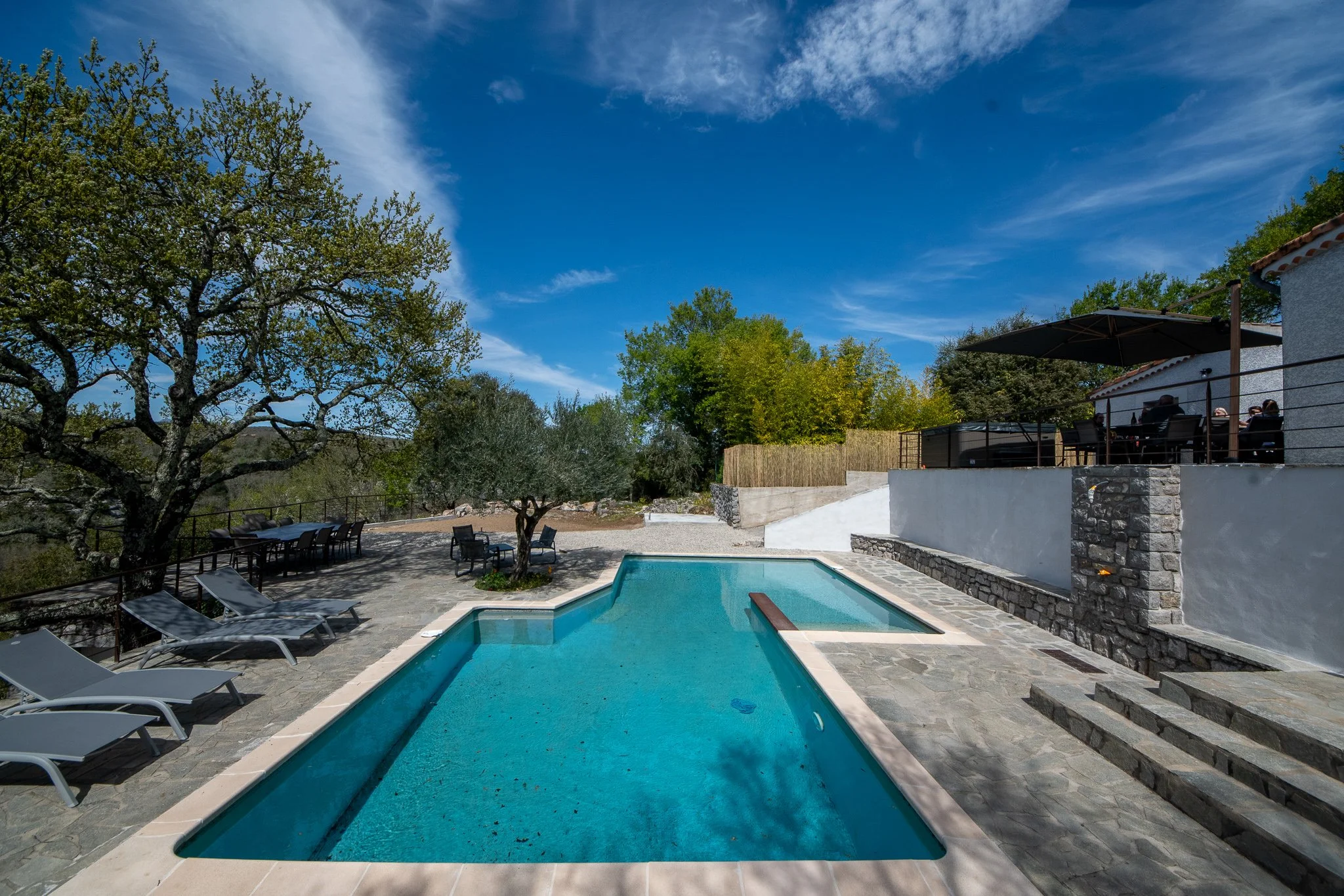 Piscine extérieure dans un espace privé avec des chaises longues, arbres et terrasse en pierre, ciel bleu avec quelques nuages, zone de détente avec mobilier et bar au fond.