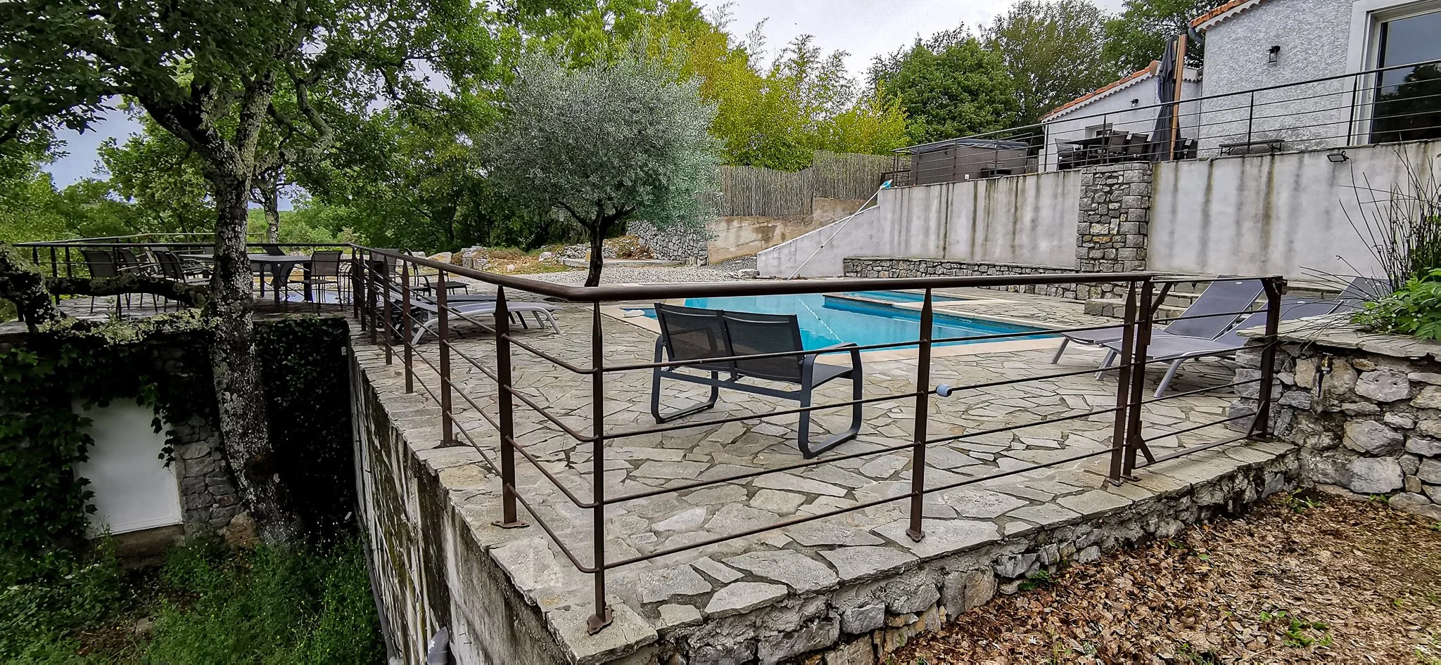 Terrasse en pierre avec piscine, chaises longues, bancs en métal, arbres et maison en arrière-plan.