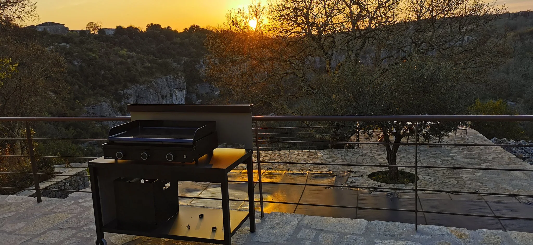 Barbecue à gaz sur une terrasse avec vue sur un coucher de soleil, arbres et paysage de montagnes.