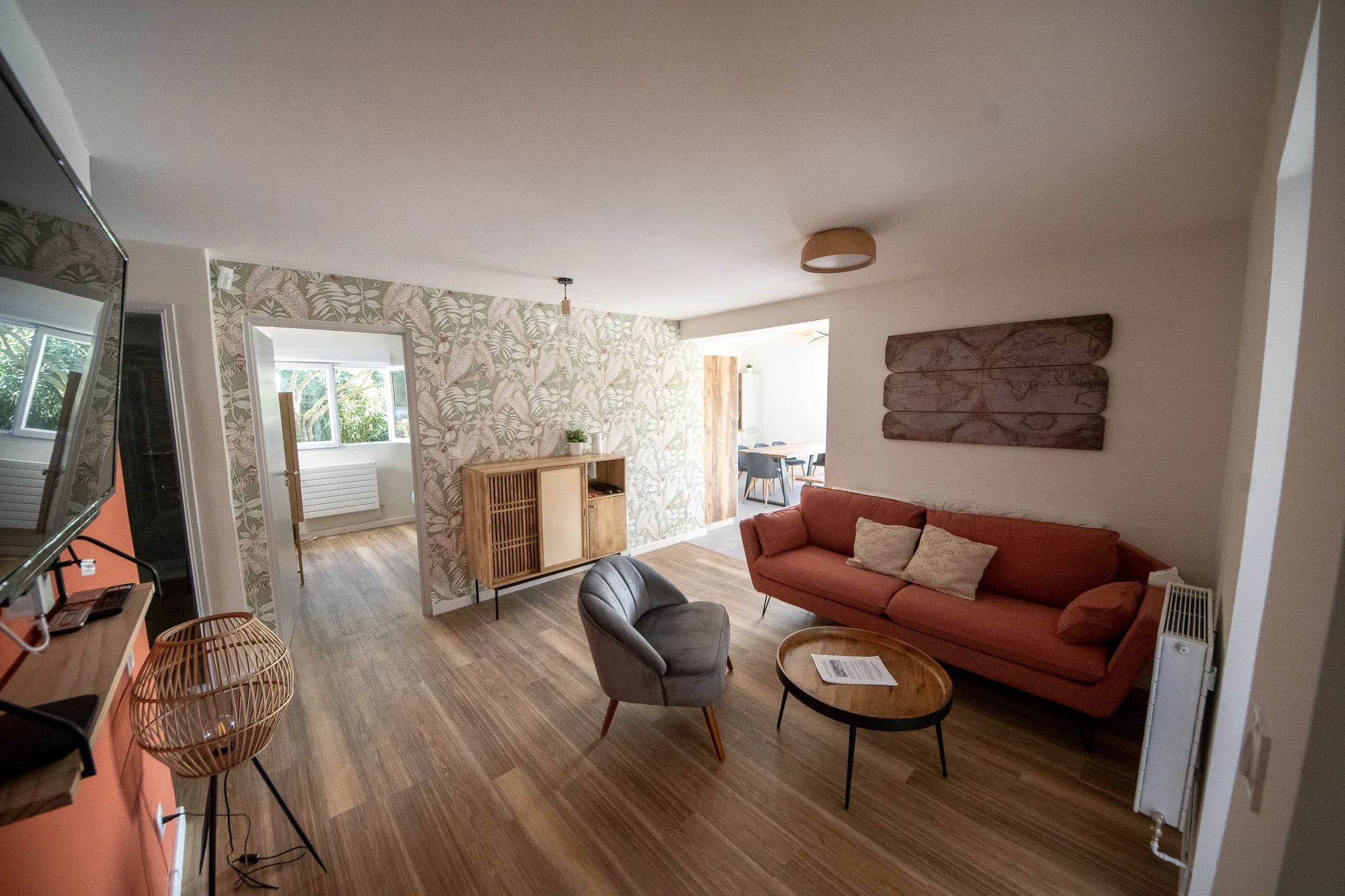 Salon moderne avec canapé orange, fauteuil gris, table basse ronde, mur avec décoration en bois, et mur en papier peint à motif tropical, fenêtres laissant entrer de la lumière naturelle.