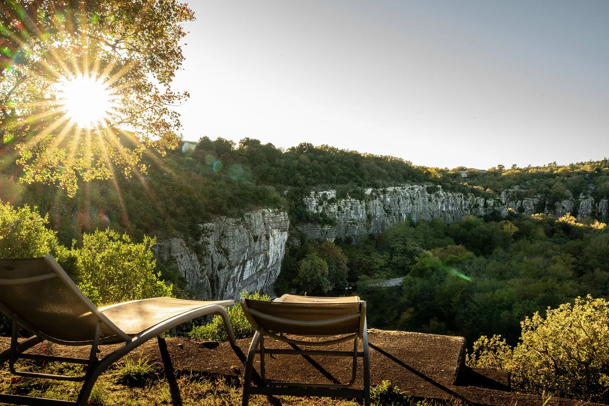 Deux chaises longues sur une terrasse avec vue sur une vallée avec des falaises caillouteuses et des arbres, avec le soleil en position bas dans le ciel.