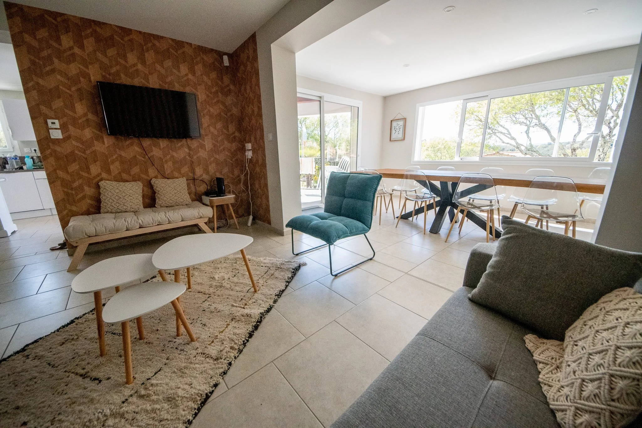 Salon lumineux avec un mur en bois, télévision murale, canapé gris, fauteuil vert, table basse blanche, tapis beige, salle à manger avec une grande table en bois et des chaises en plastique transparent, grande fenêtre offrant une vue sur la nature.