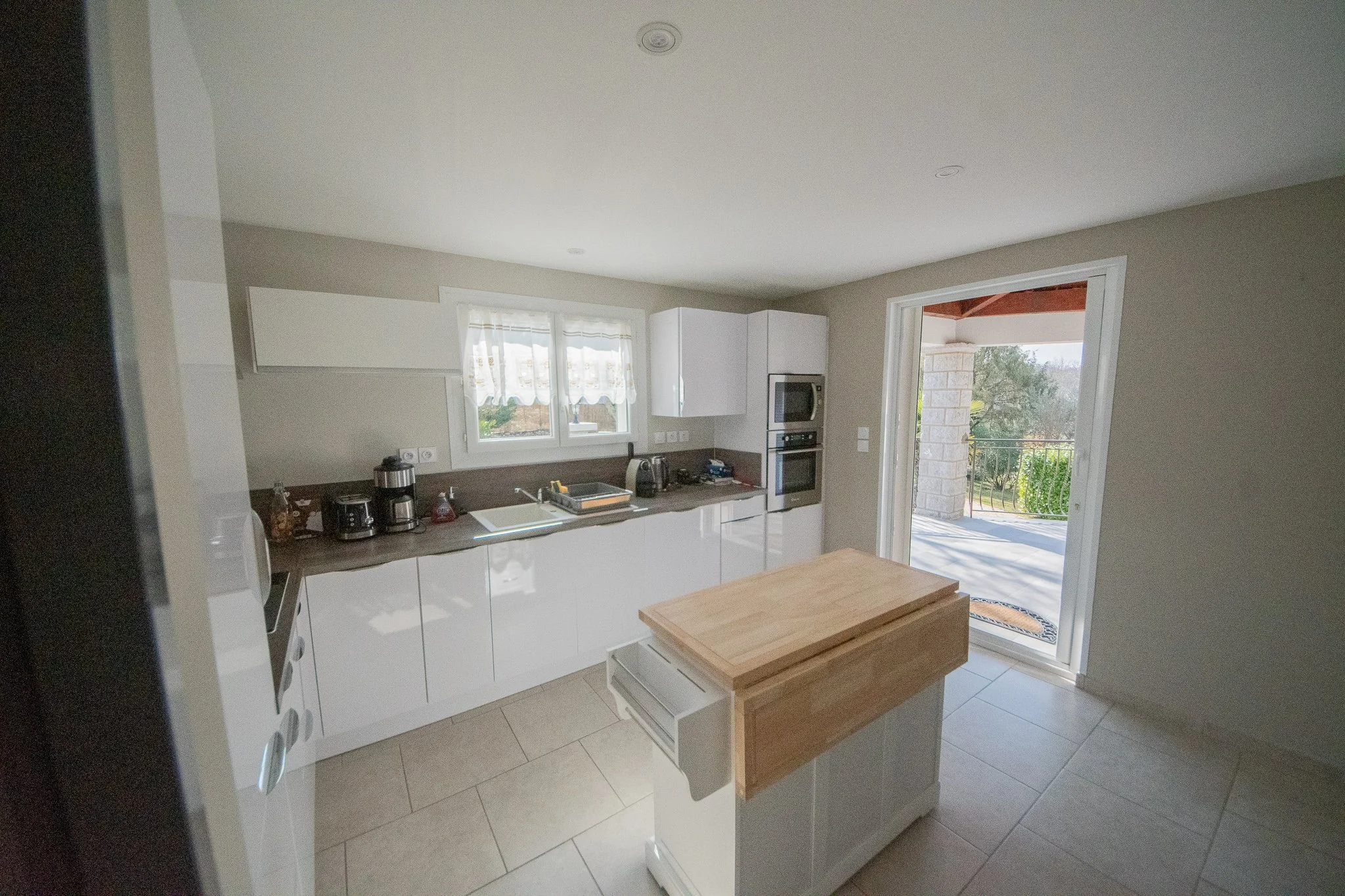 Cuisine moderne avec comptoir en bois, électroménagers blancs, une porte-fenêtre donnant sur une terrasse extérieure en pierre et bois, vue sur un jardin