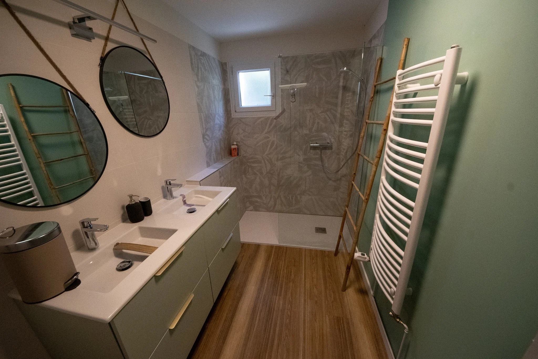 Salle de bain avec lavabo double, miroir rond, douche à l'italienne, étendoir en bois, radiateur porte-serviettes blanc