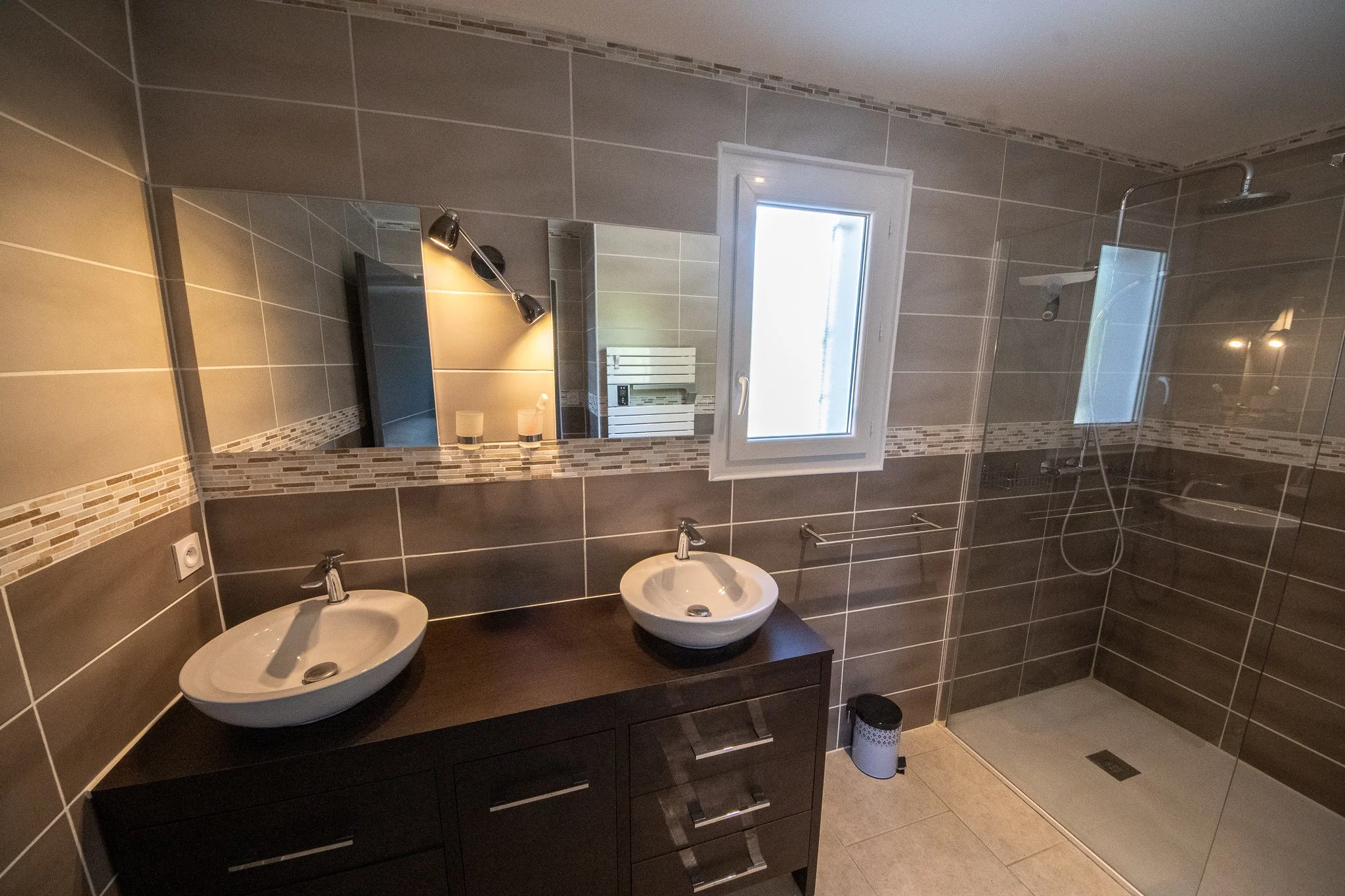 Salle de bain avec deux lavabos ovales sur un meuble en bois foncé, grand miroir mural, petites mosaïques décoratives, fenêtre laissant entrer la lumière, douche avec paroi en verre et pomme de douche, évier, accessoire de salle de bain, petite poube