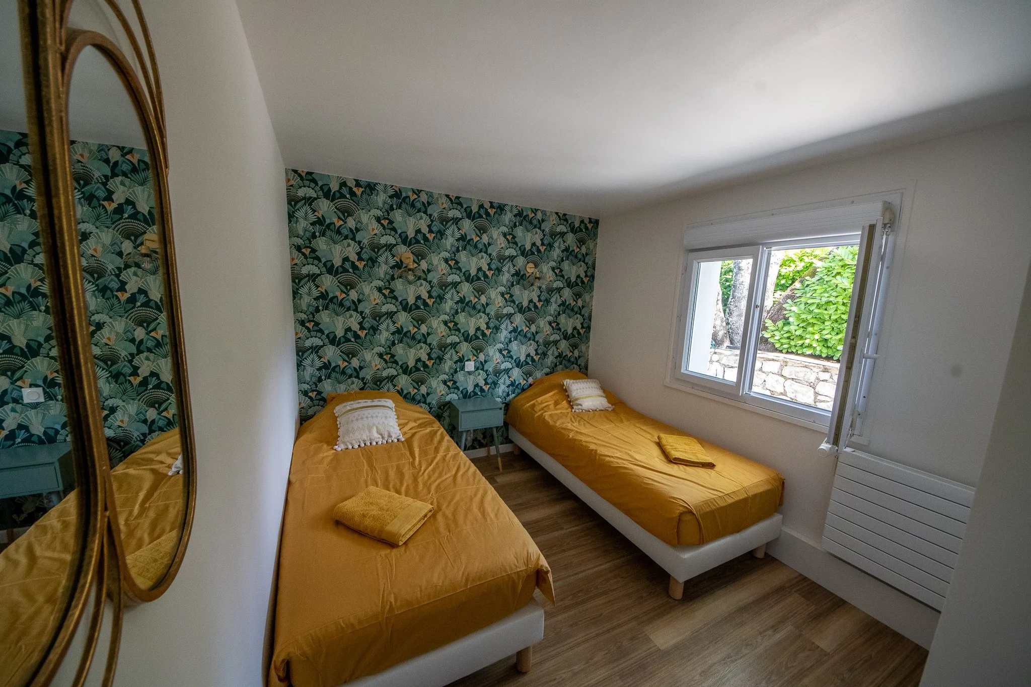 Chambre avec deux lits simples avec draps jaunes, un mur avec papier peint à motif végétal, fenêtre laissant entrer la lumière naturelle, miroir ancien à gauche.