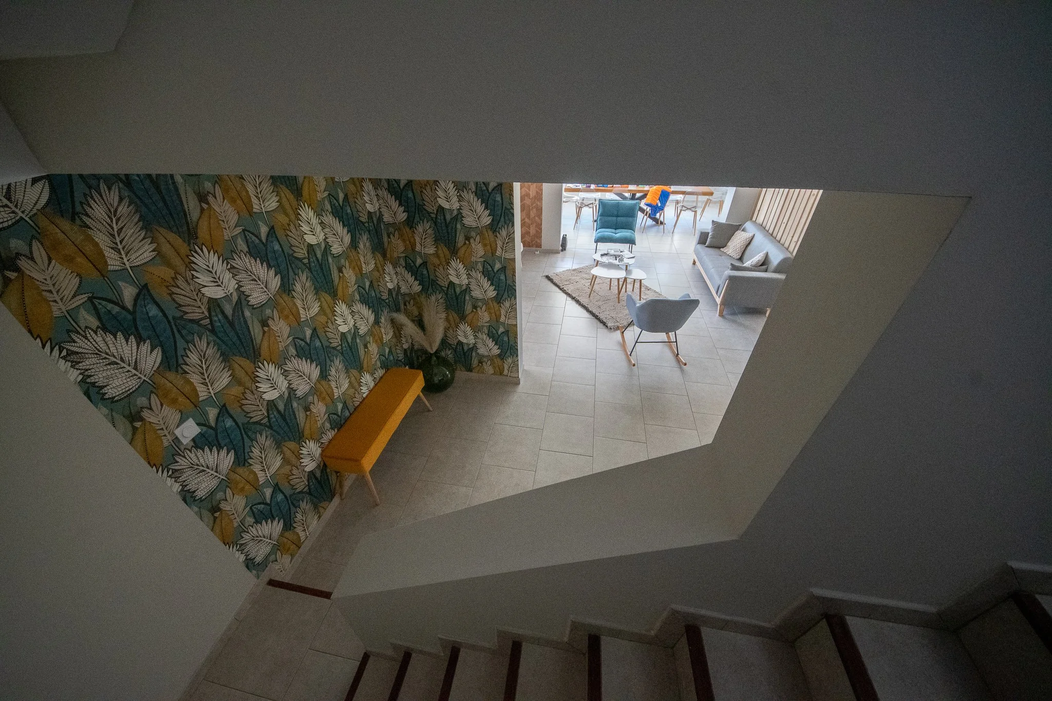 Vue en plongée d'un salon moderne avec un mur à motifs feuillage, un banc orange, un coussin gris, un tapis beige, plusieurs fauteuils et canapés gris, une table basse, et une lumière naturelle.