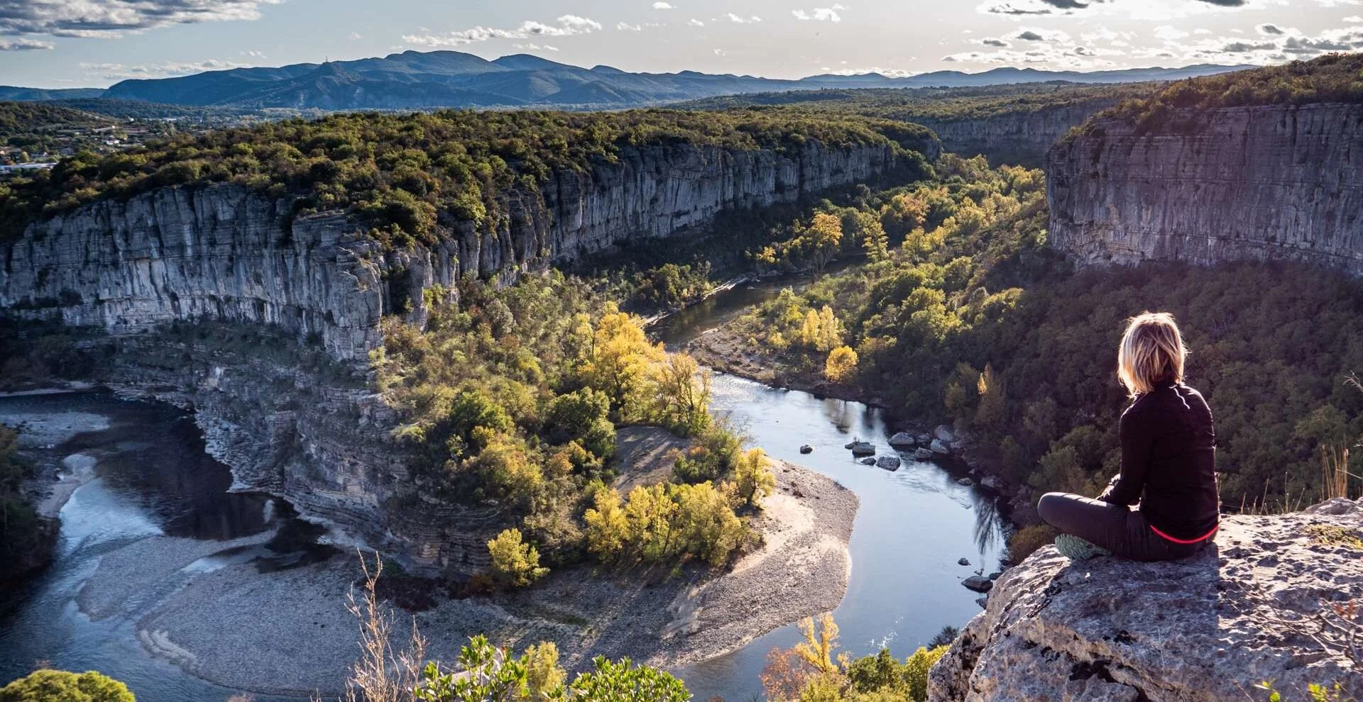 Une personne assise sur un rocher, Cirque de Gens, Chauzon, Ardèche, observant une vallée avec une rivière serpentant entre des falaises rocheuses verdoyantes, sous un ciel partiellement nuageux en fin d'après-midi