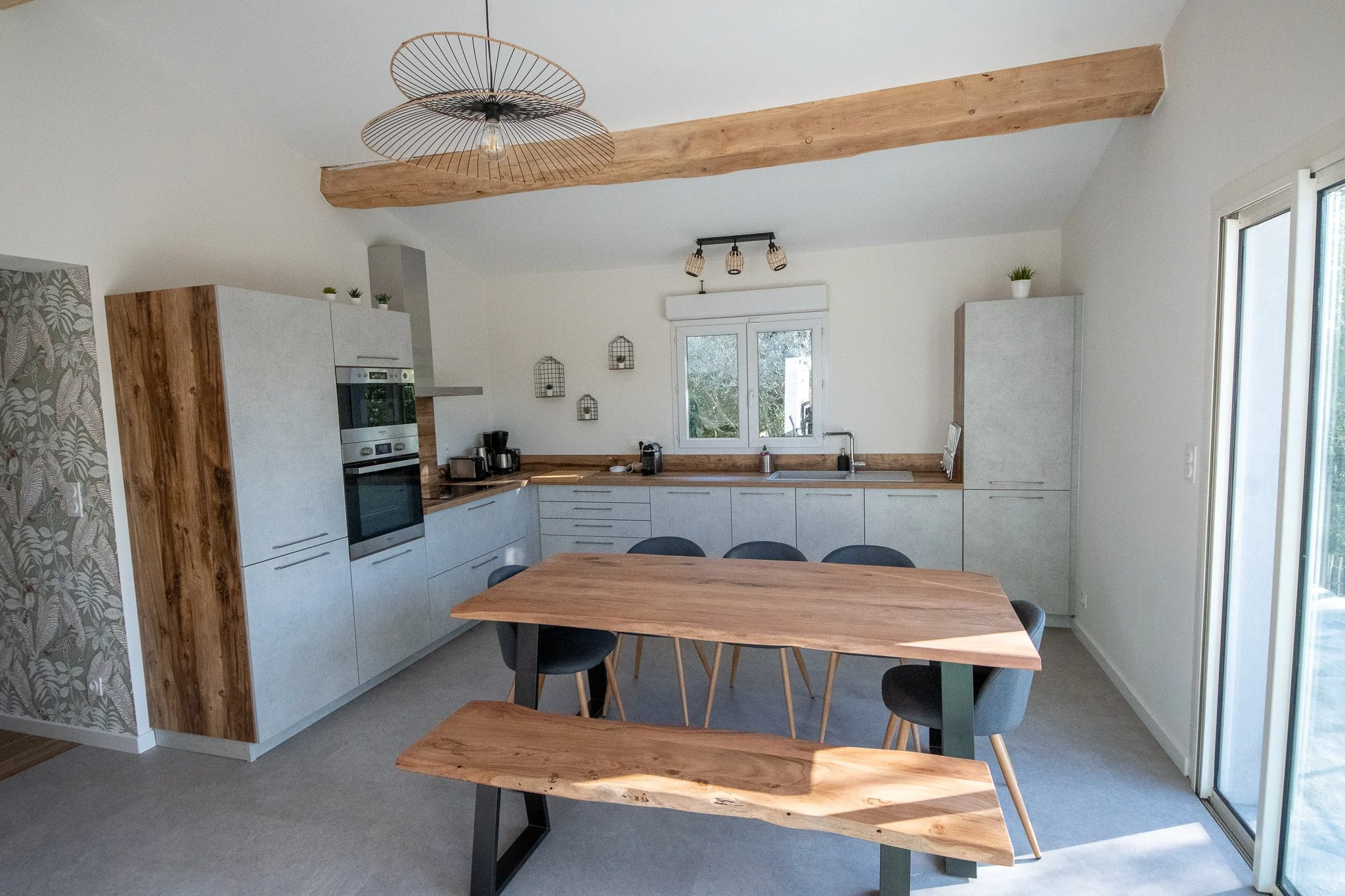 Cuisine moderne avec meubles en bois clair et blanc, table en bois, chaises en gris foncé, fenêtre et porte-fenêtre qui donnent sur l'extérieur, éclairage suspendu et décorations murales minimalistes.