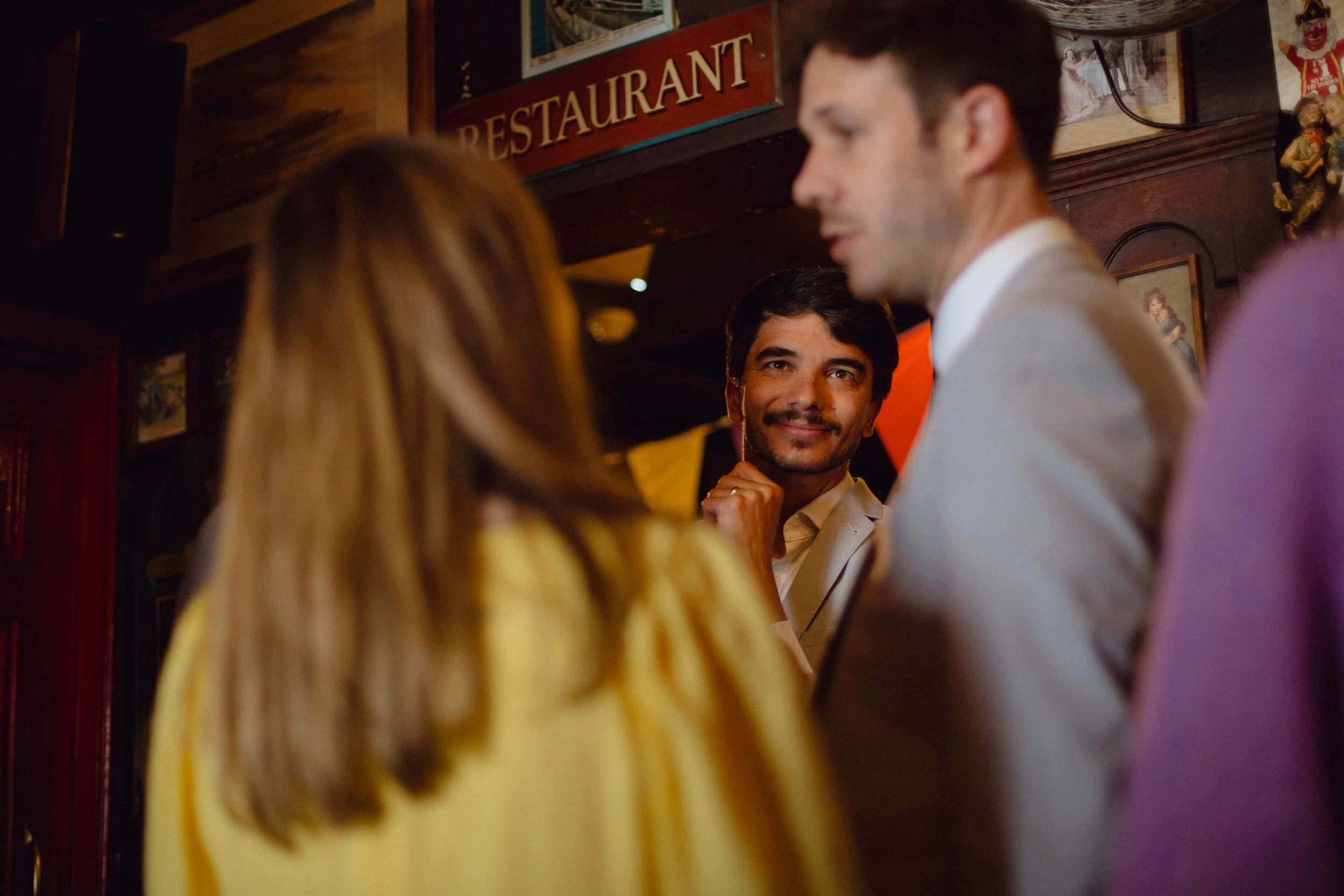 A group of four people engaged in conversation inside a restaurant, with a man smiling and looking at a woman with brown hair, and a man with dark hair and a beard as well as a woman with long blonde hair facing away from the camera.
