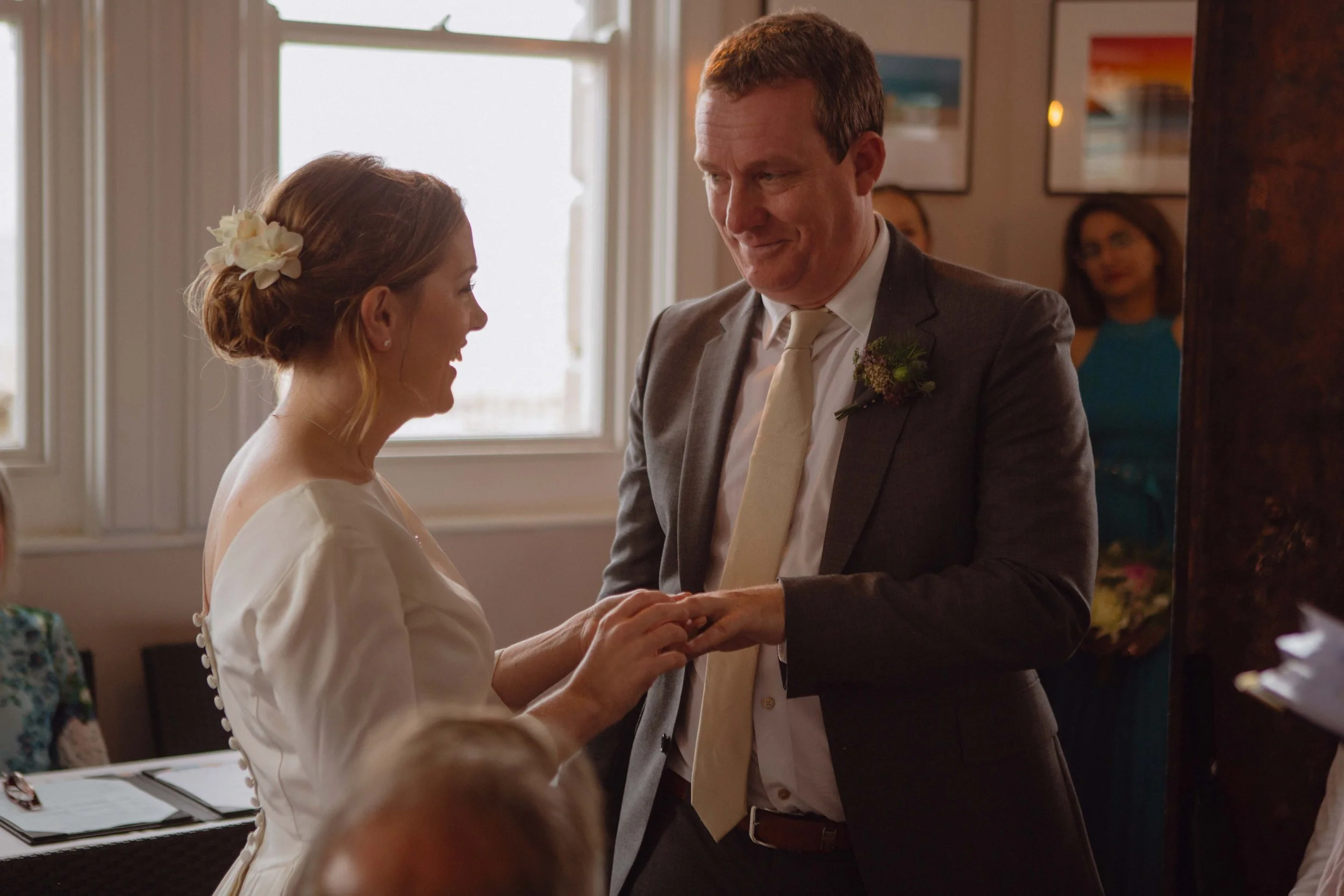 A bride and groom exchanging rings during a wedding ceremony inside a room with large windows and framed pictures on the wall.