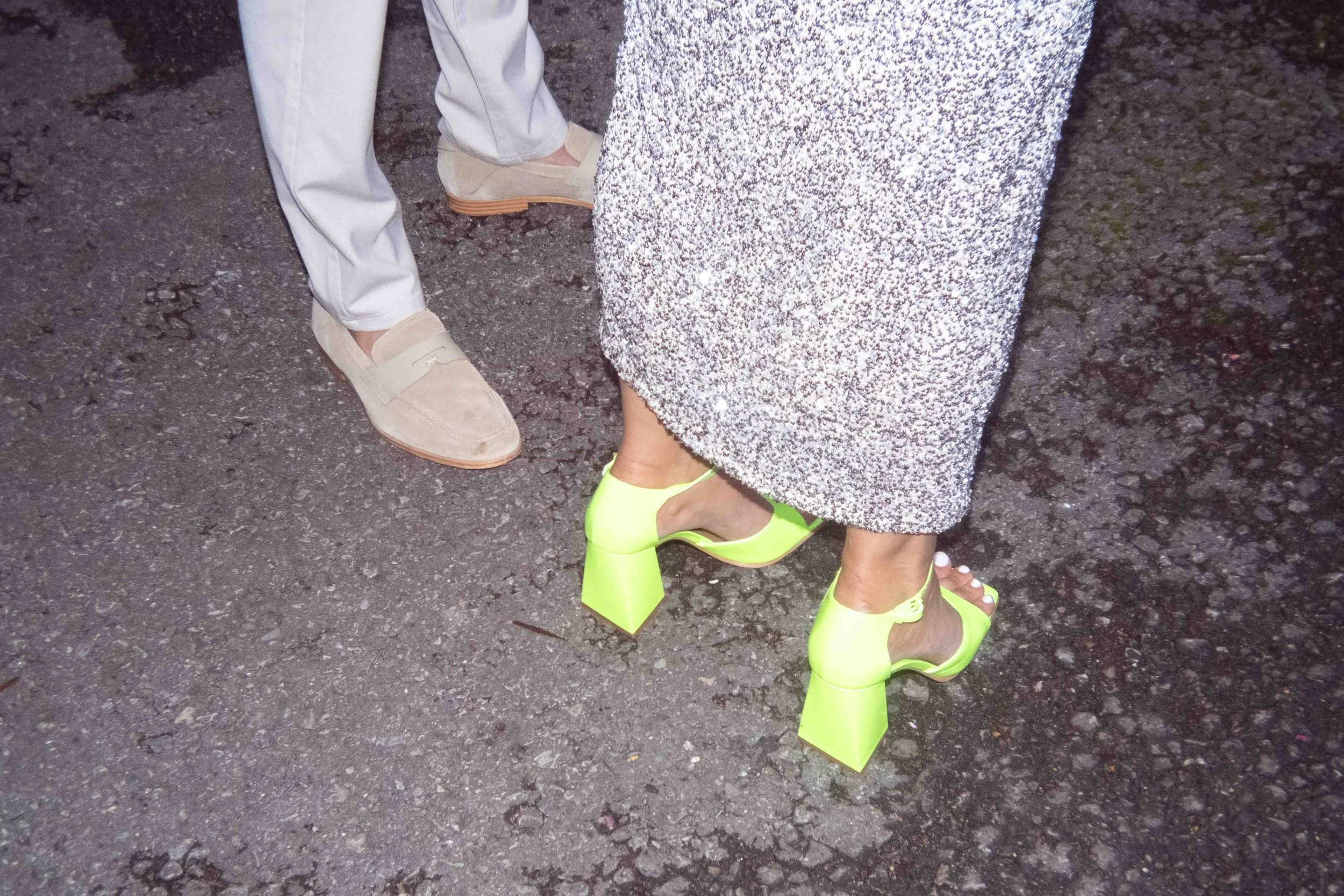 Close-up of a woman's feet wearing bright yellow high-heeled shoes with chunky square heels, standing on a textured asphalt ground, next to a man wearing beige loafers and light-colored pants.