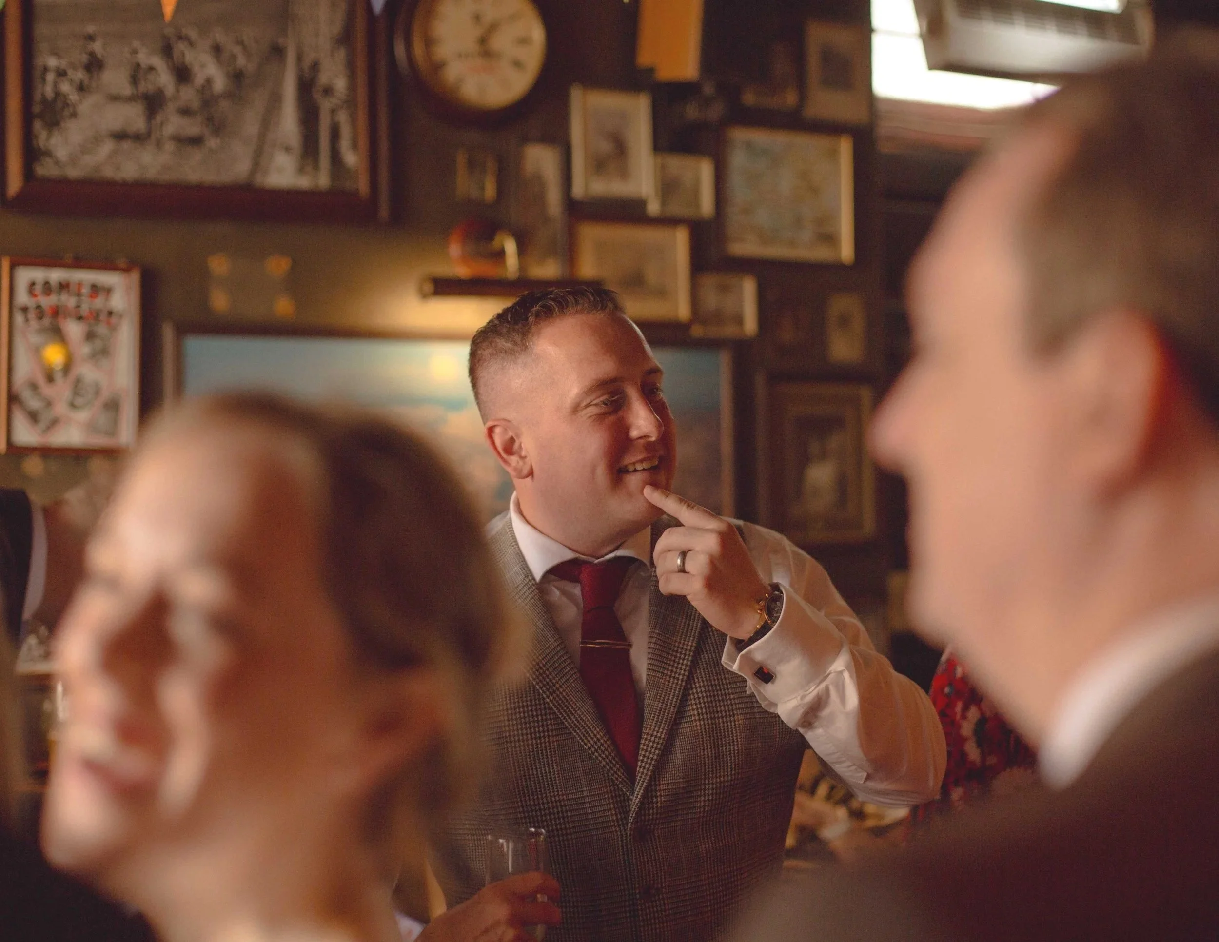 A man in a plaid suit and red tie smiling and touching his chin while talking to people at a social gathering in a bar or restaurant with framed pictures and a clock on the wall.