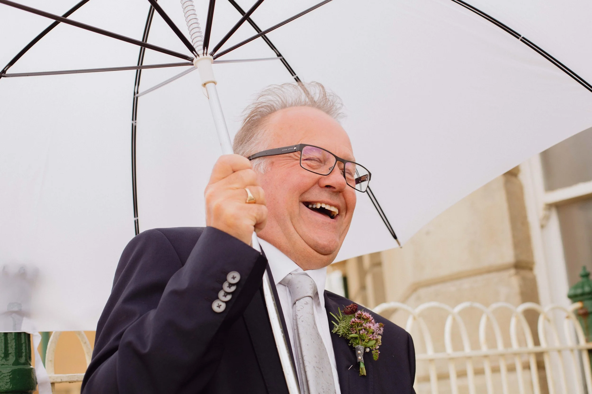 Smiling man in a tuxedo, wearing glasses and a boutonniere, holding an umbrella outdoors.