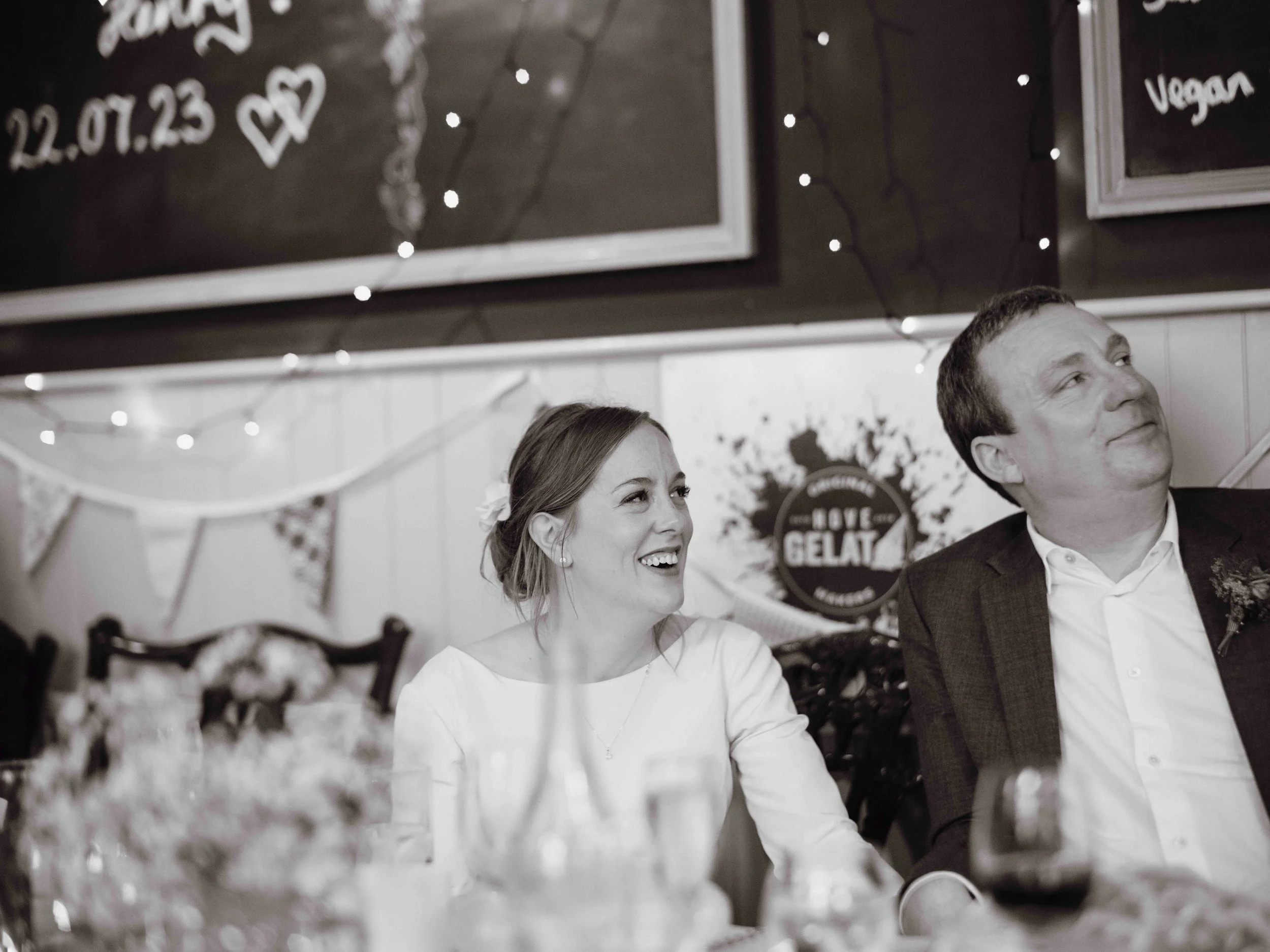A woman and a man sitting at a decorated celebration table, smiling and looking to the right. The woman has light hair styled back with a flower accessory, and the man is wearing a dark blazer over a white shirt. The background has festive string lig