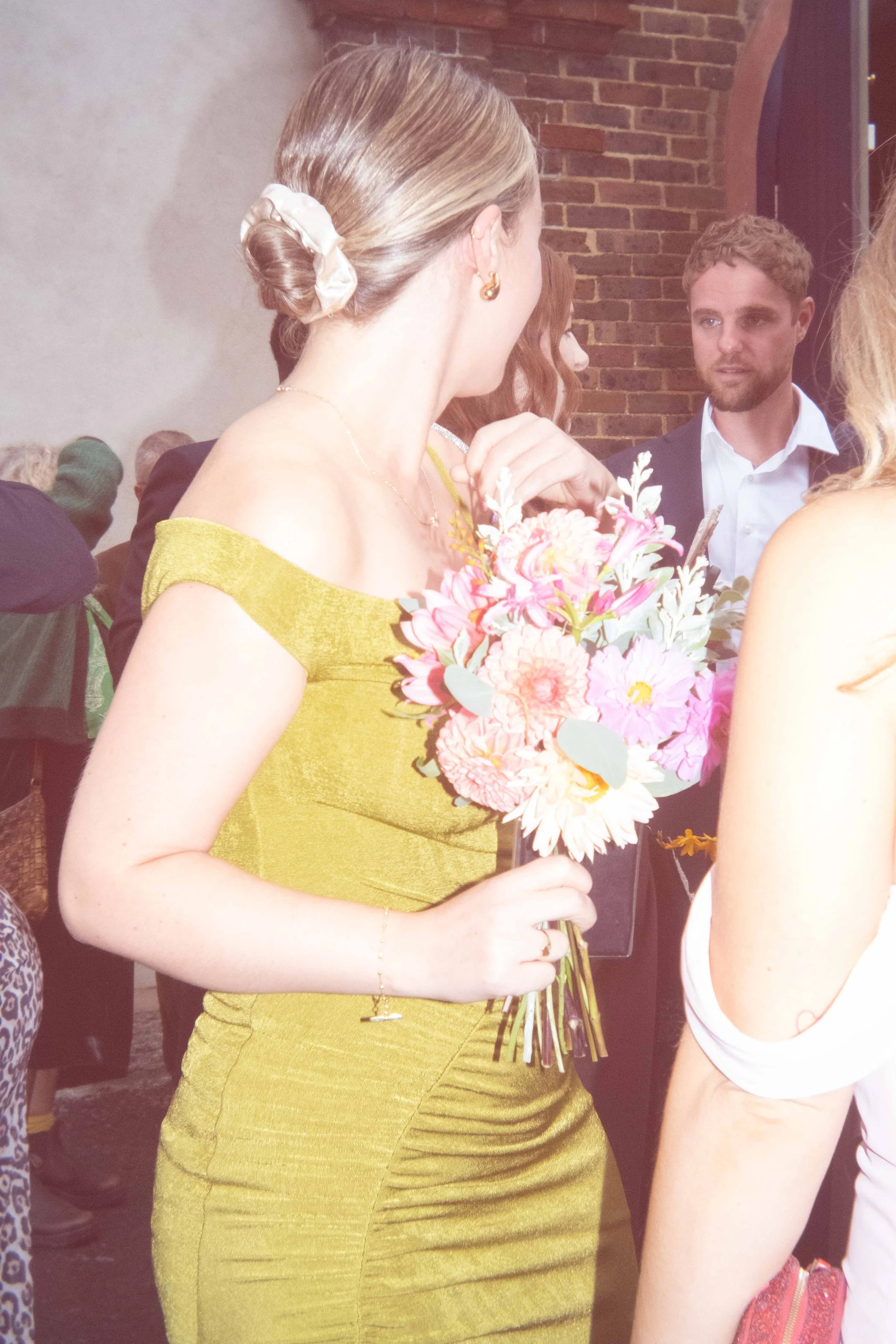 A woman in a yellow dress holding a bouquet of pink flowers at a social event or gathering.