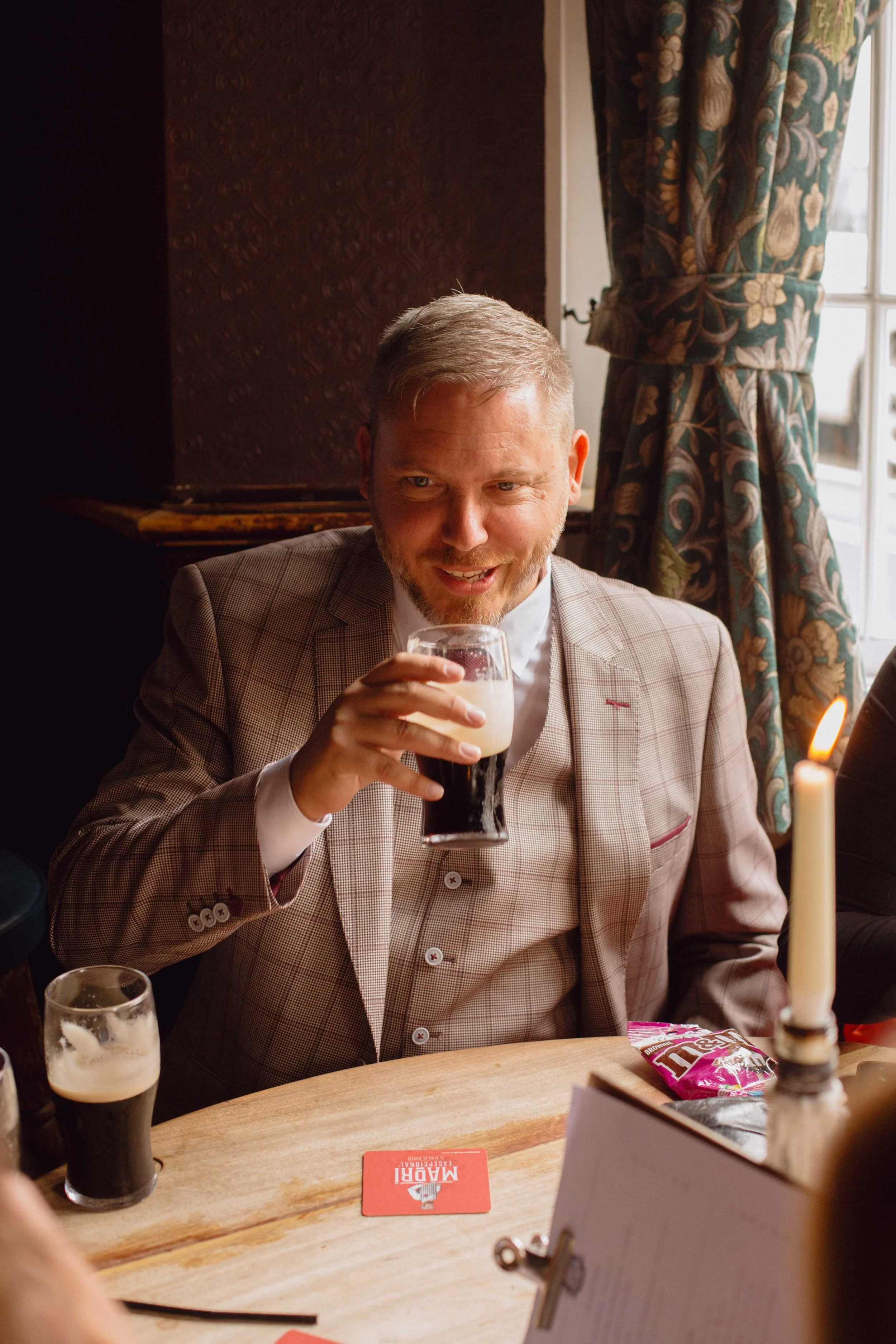 A man in a checkered blazer sitting at a table, holding a glass of dark beer with a foam head, smiling, with candles and snacks on the table inside a cozy, decorated room.