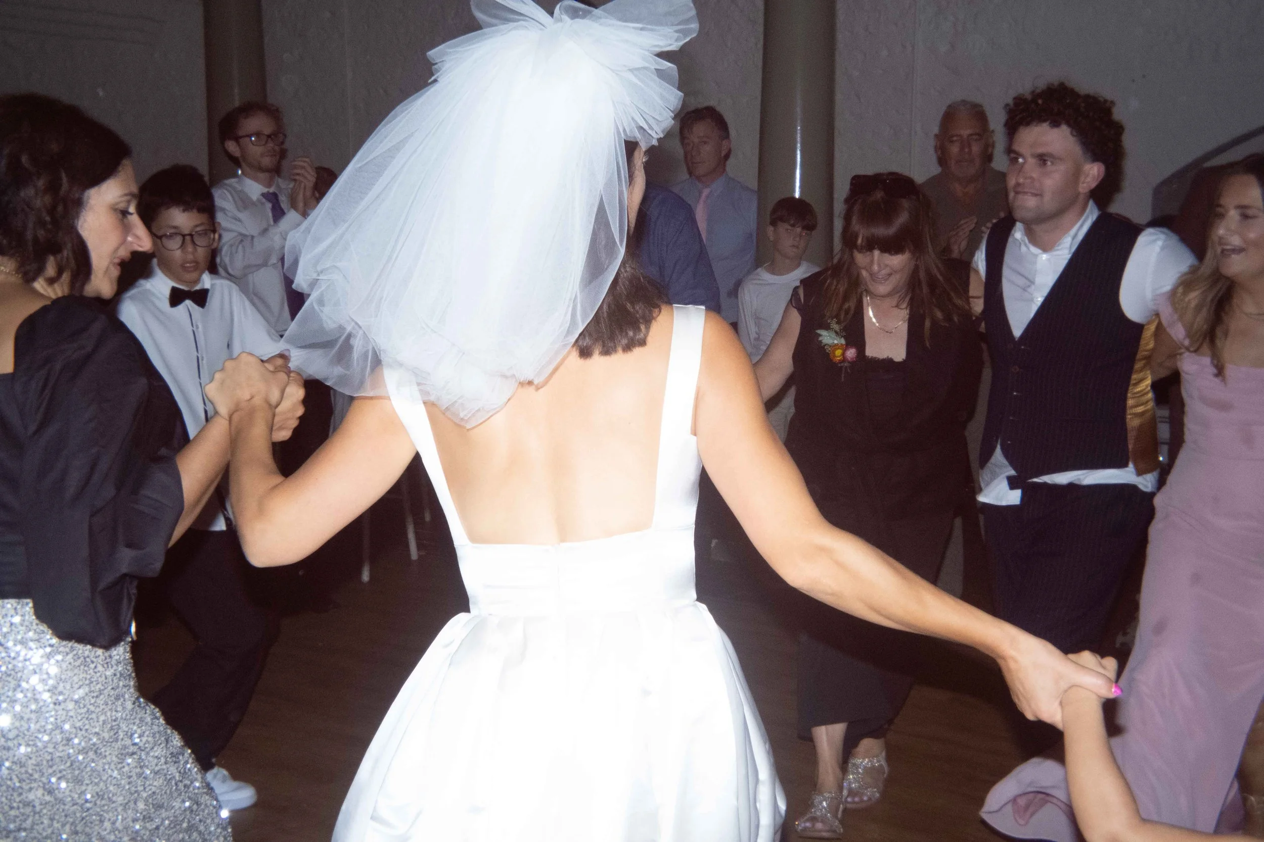 People dancing in a circle at a wedding reception, with the bride in a white dress and veil in the center.
