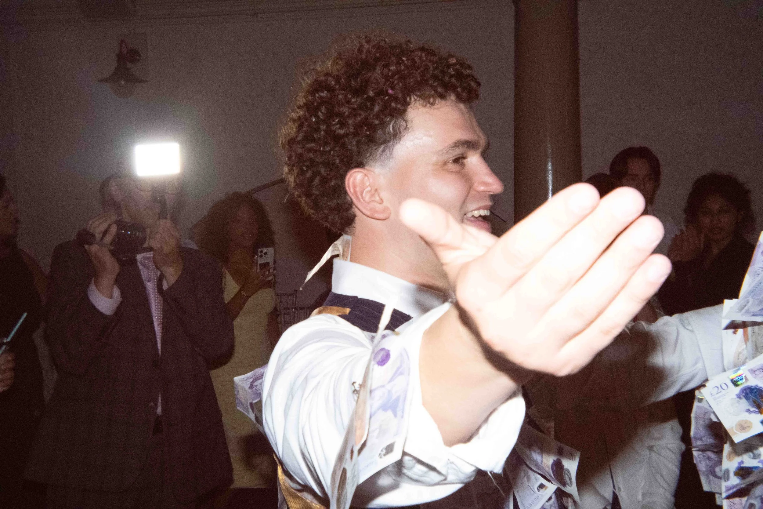 A man with curly hair smiling and raising his hand at a celebration or event, surrounded by people taking photos and holding money.