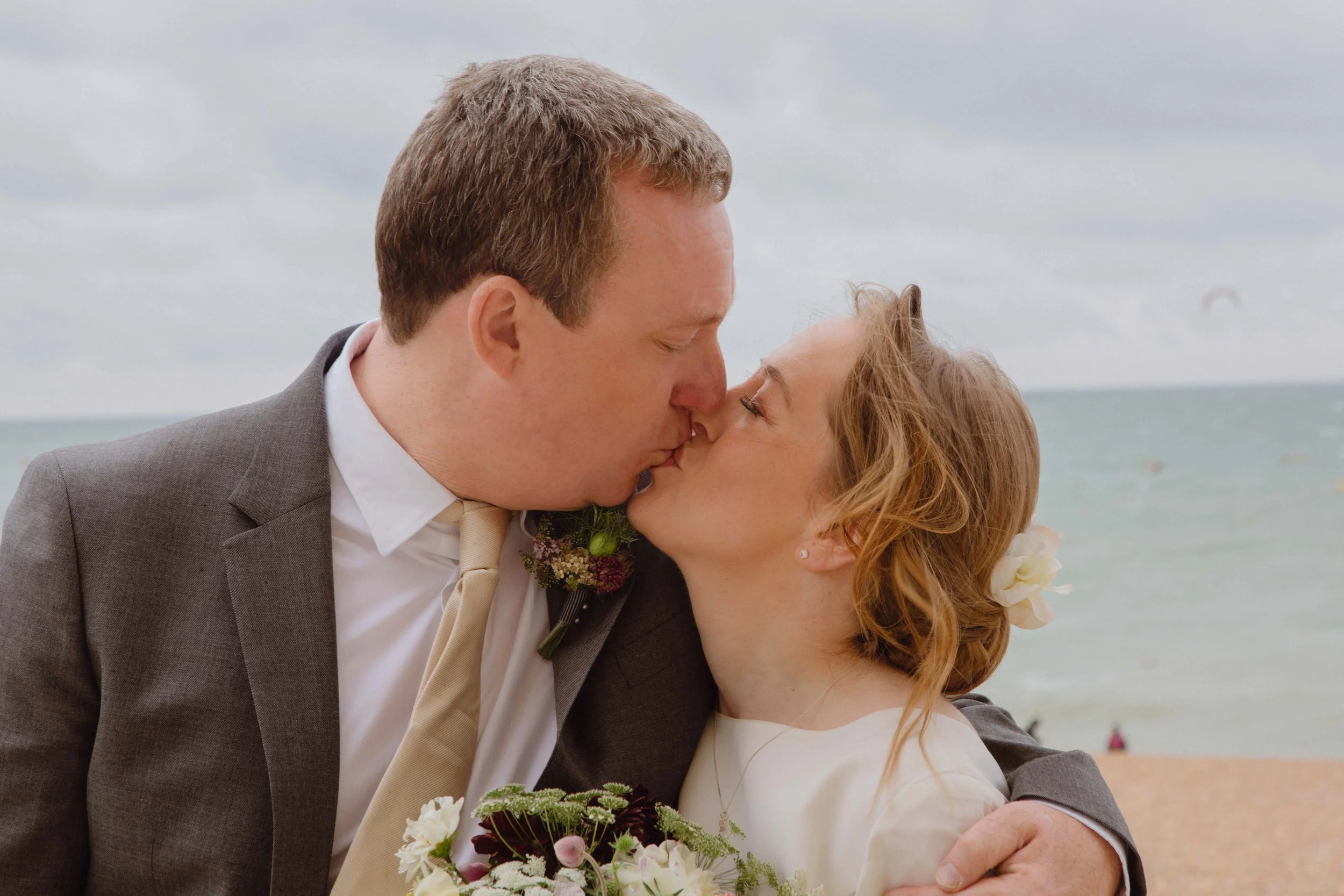 A couple sharing a kiss at a beach wedding, with the ocean and cloudy sky in the background.