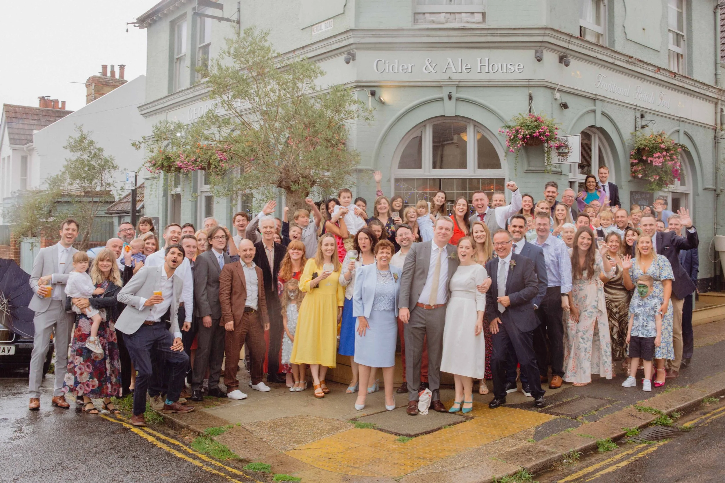 Large group of people gathered outside a building with a sign that reads 'Cider & Ale House,' celebrating a special occasion, with some raising hands and holding drinks, on a rainy day.