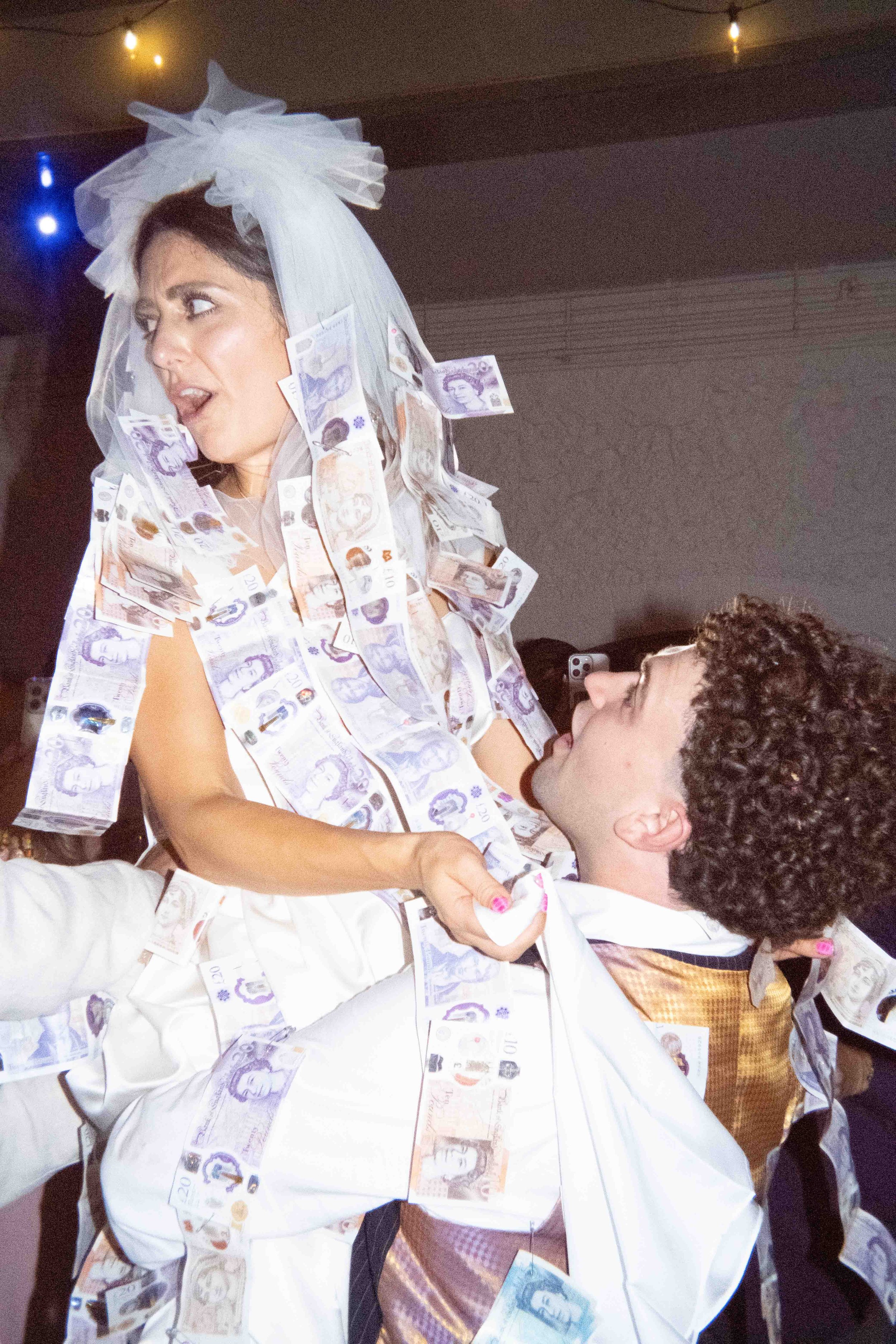A woman dressed in white is covered in hanging and scattered £20 and £10 banknotes, with a woman looking up at her, in a dimly lit room.