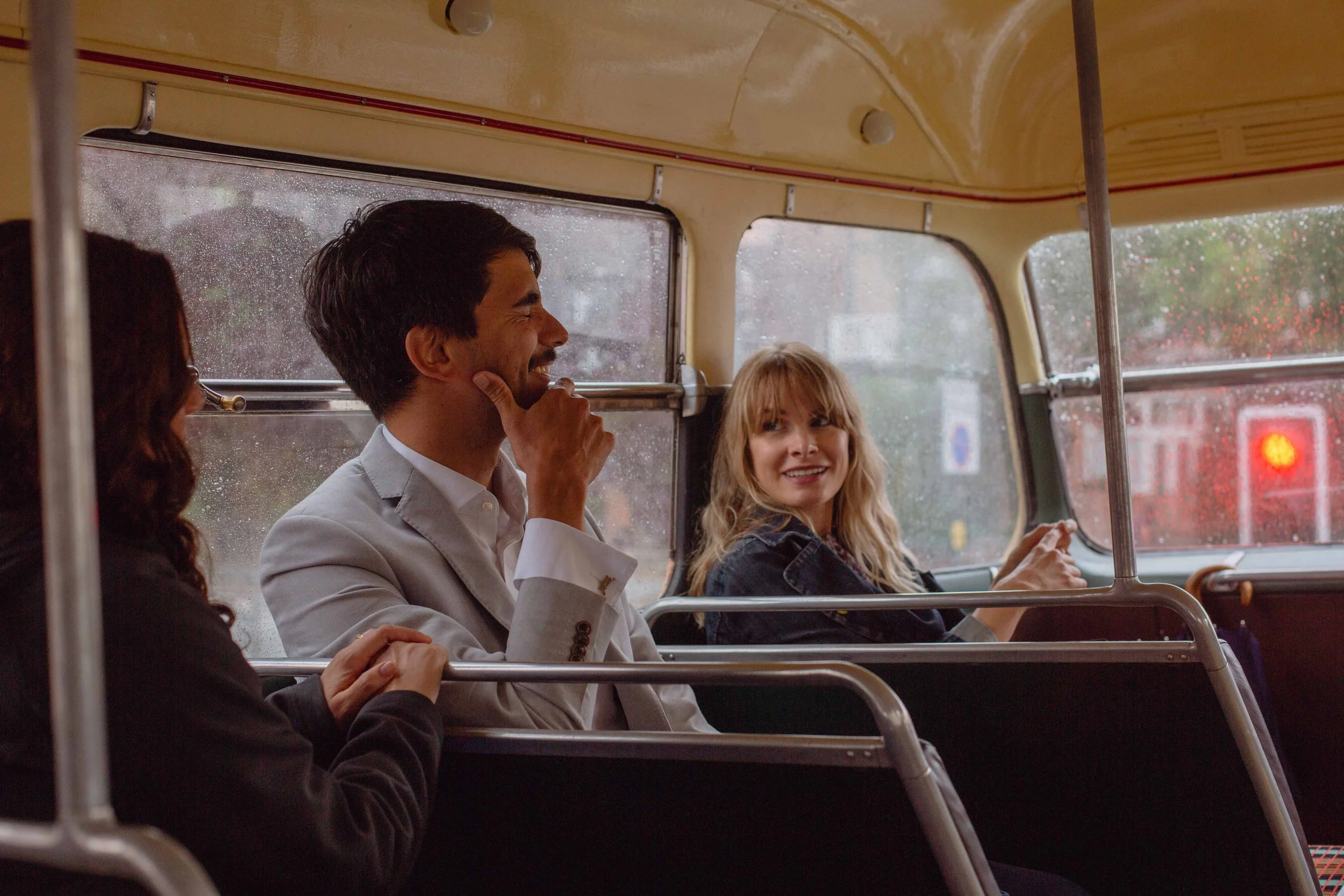 Three people sitting on a bus, smiling and chatting. The rain is visible outside the window.