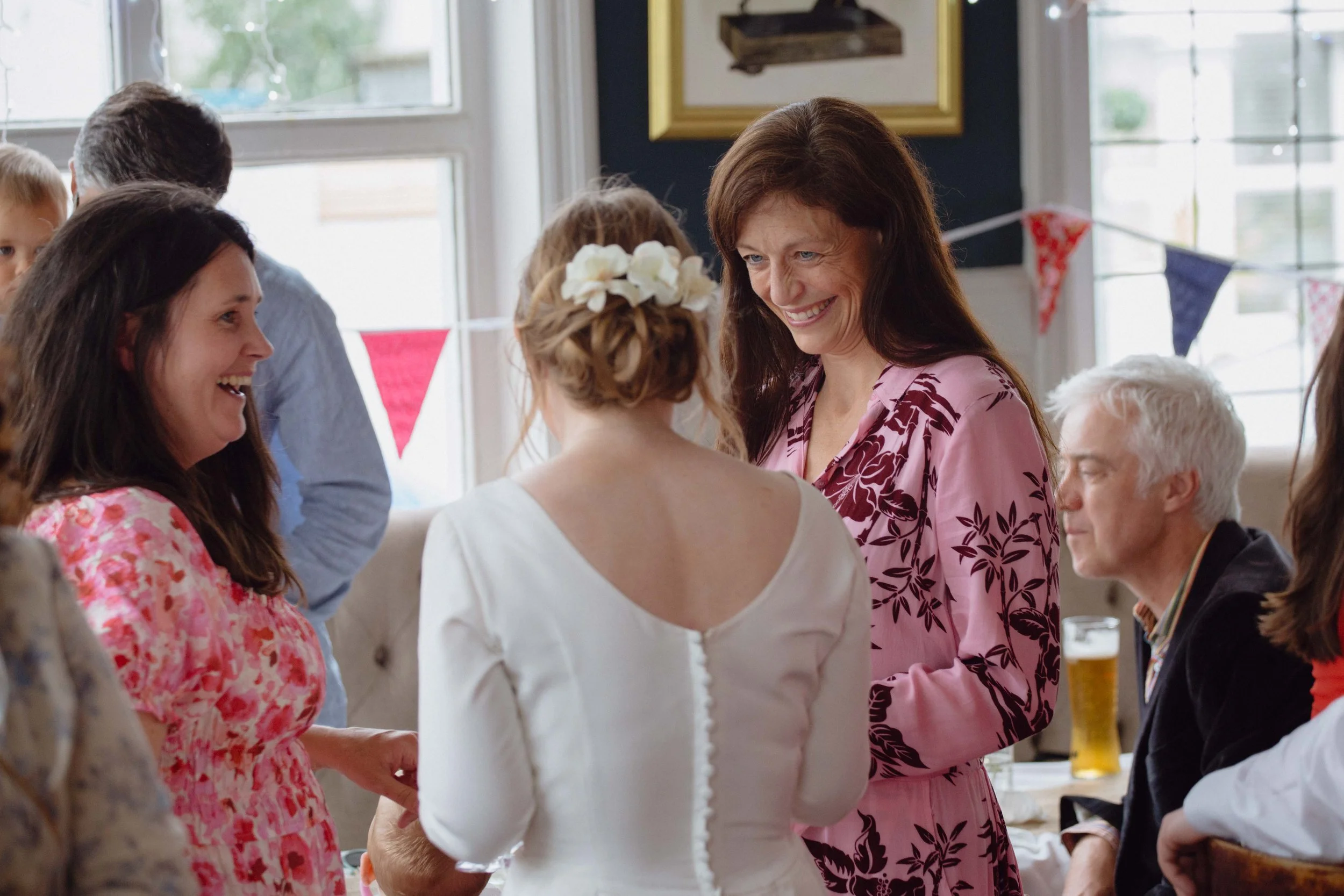 A woman in a pink floral dress smiling in conversation with a woman in a white dress with a floral headpiece at a celebration or gathering indoors, with multicolored bunting in the background.