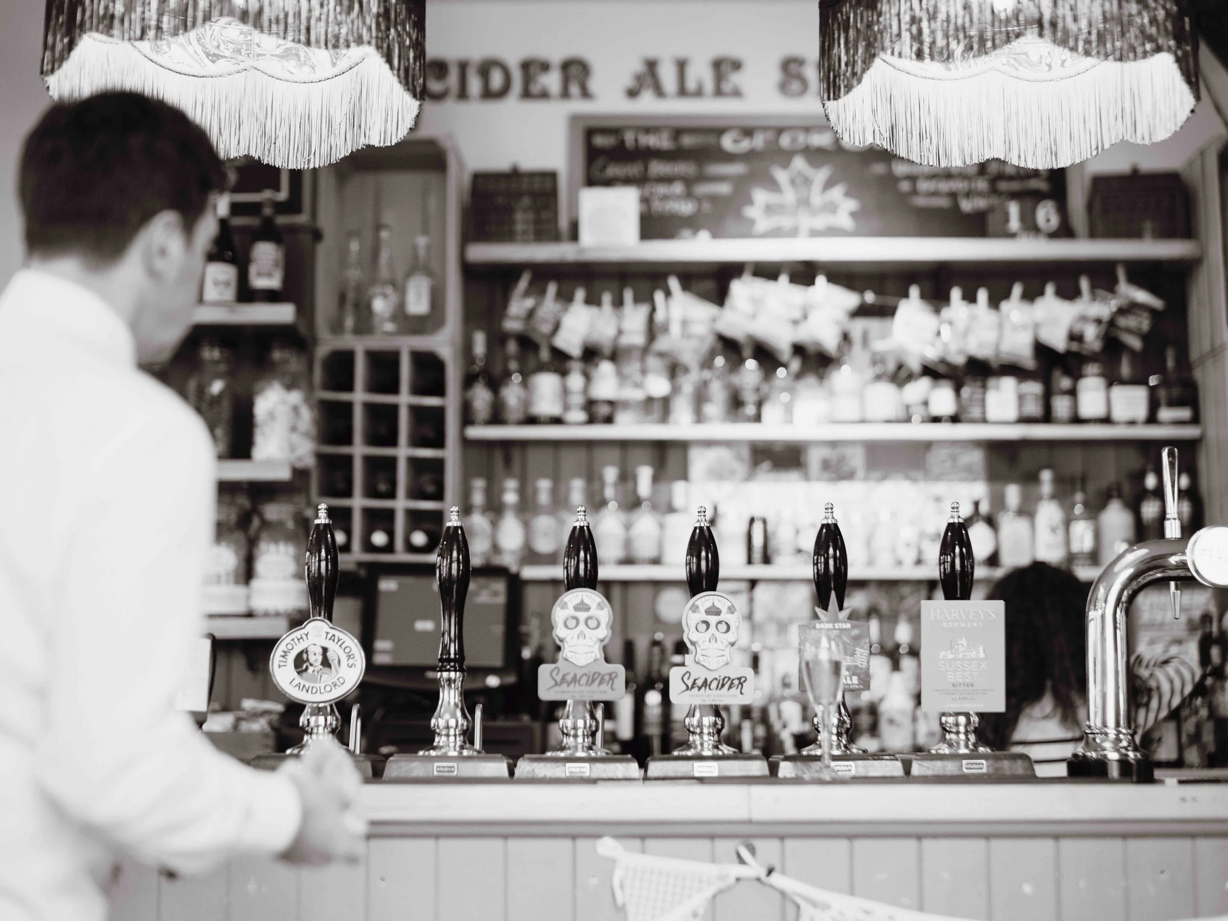 Black and white photo of a bartender or server behind a bar counter with beer taps, a man in white shirt in the foreground, shelves with bottles and snacks behind, and hanging lamps overhead.