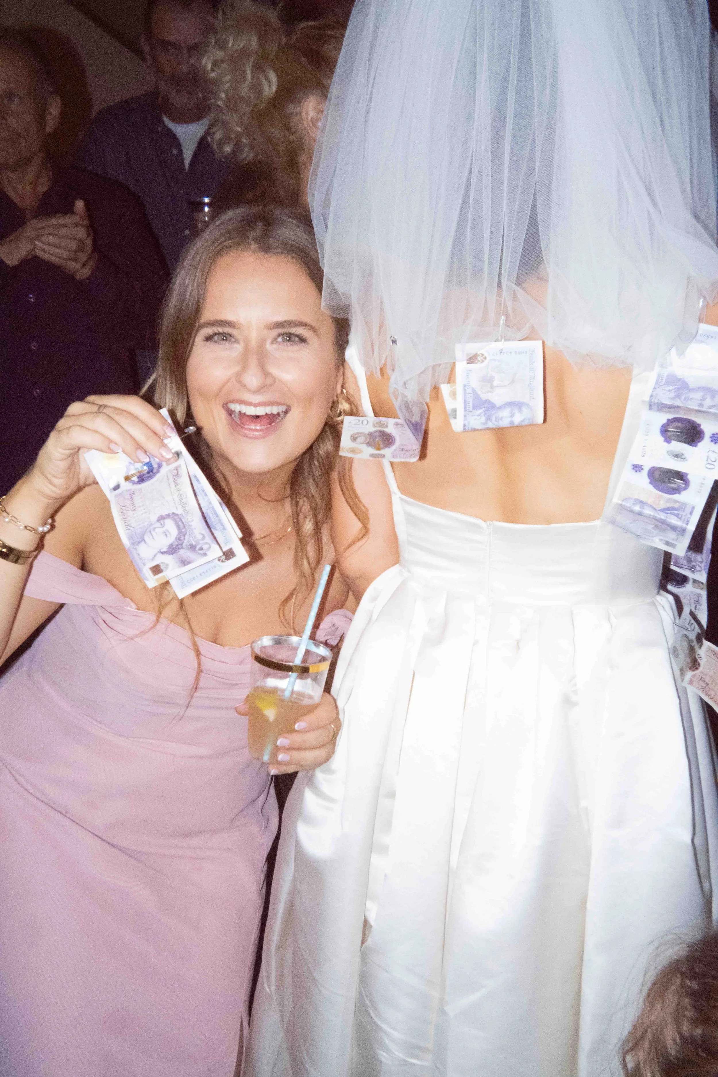 Woman holding a 20-pound note and a drink, smiling at a party with another person dressed in a white dress and veil with money pinned to it, in a dimly lit room with other people in the background.