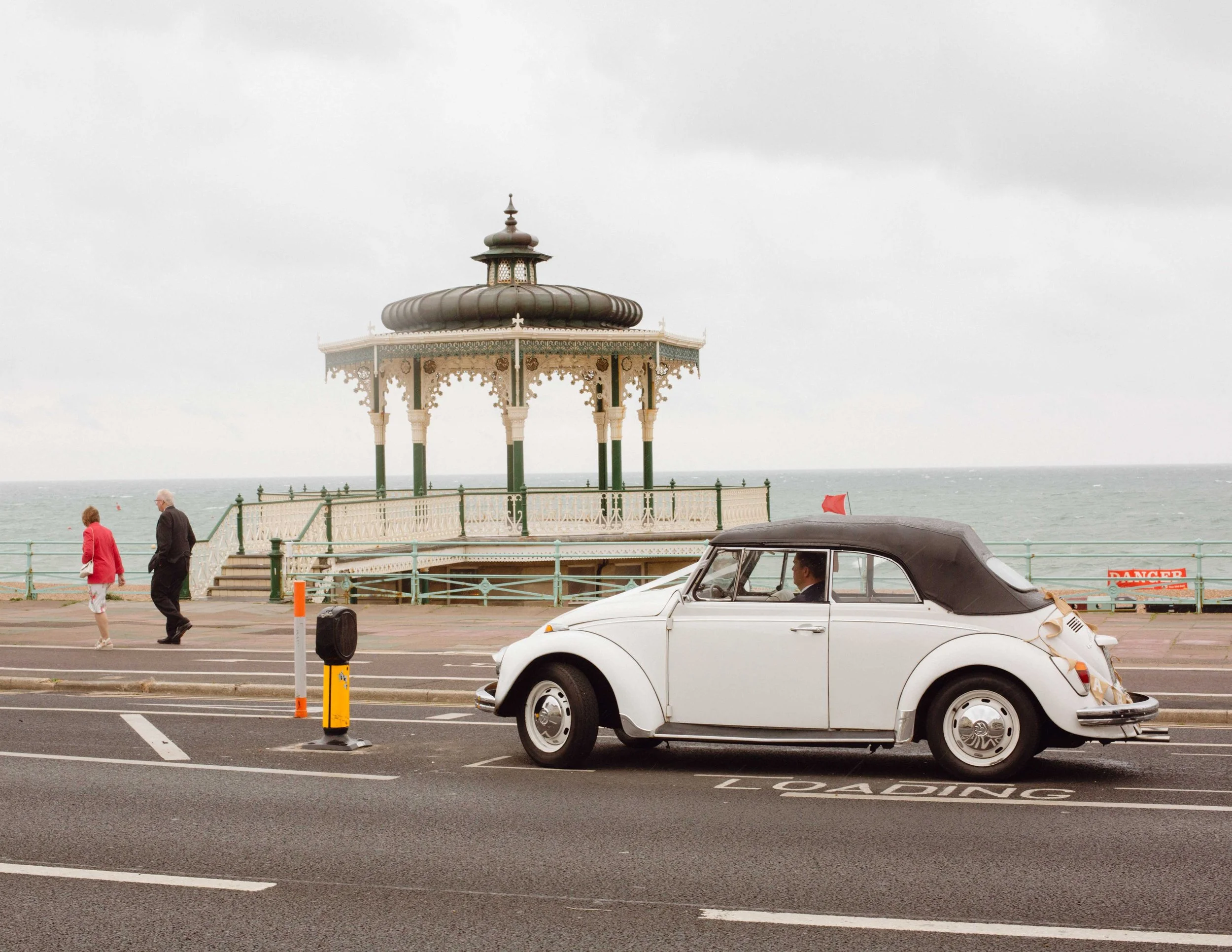A vintage white convertible car parked in a parking space near a seaside promenade with a Victorian-style gazebo structure overlooking the ocean. Two people walk along the promenade, and a red flag is visible in the background.