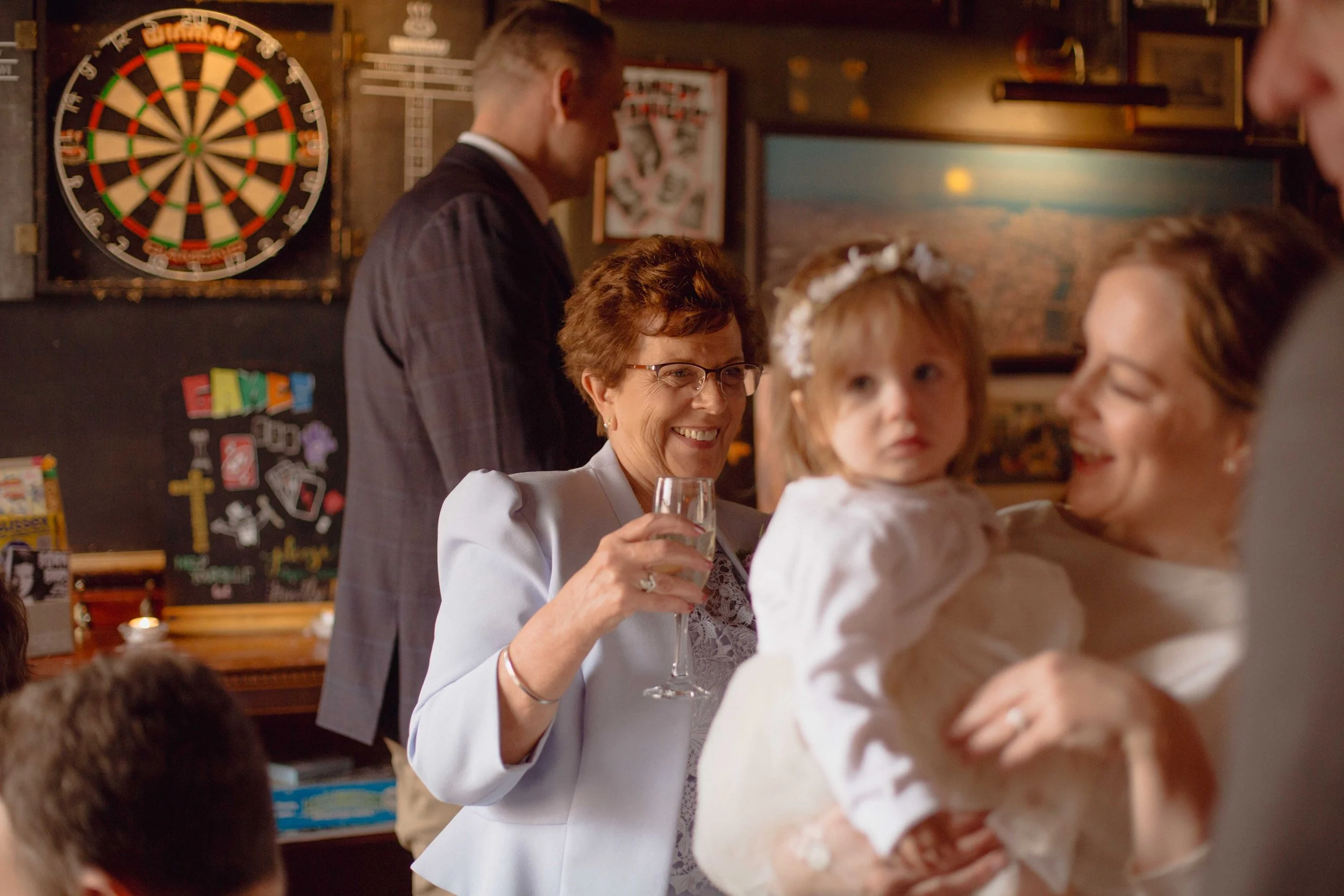 A woman holding a young girl at a celebration, with a dartboard on the wall and a chalkboard with colorful magnets behind them. The woman is smiling and holding a glass of champagne.