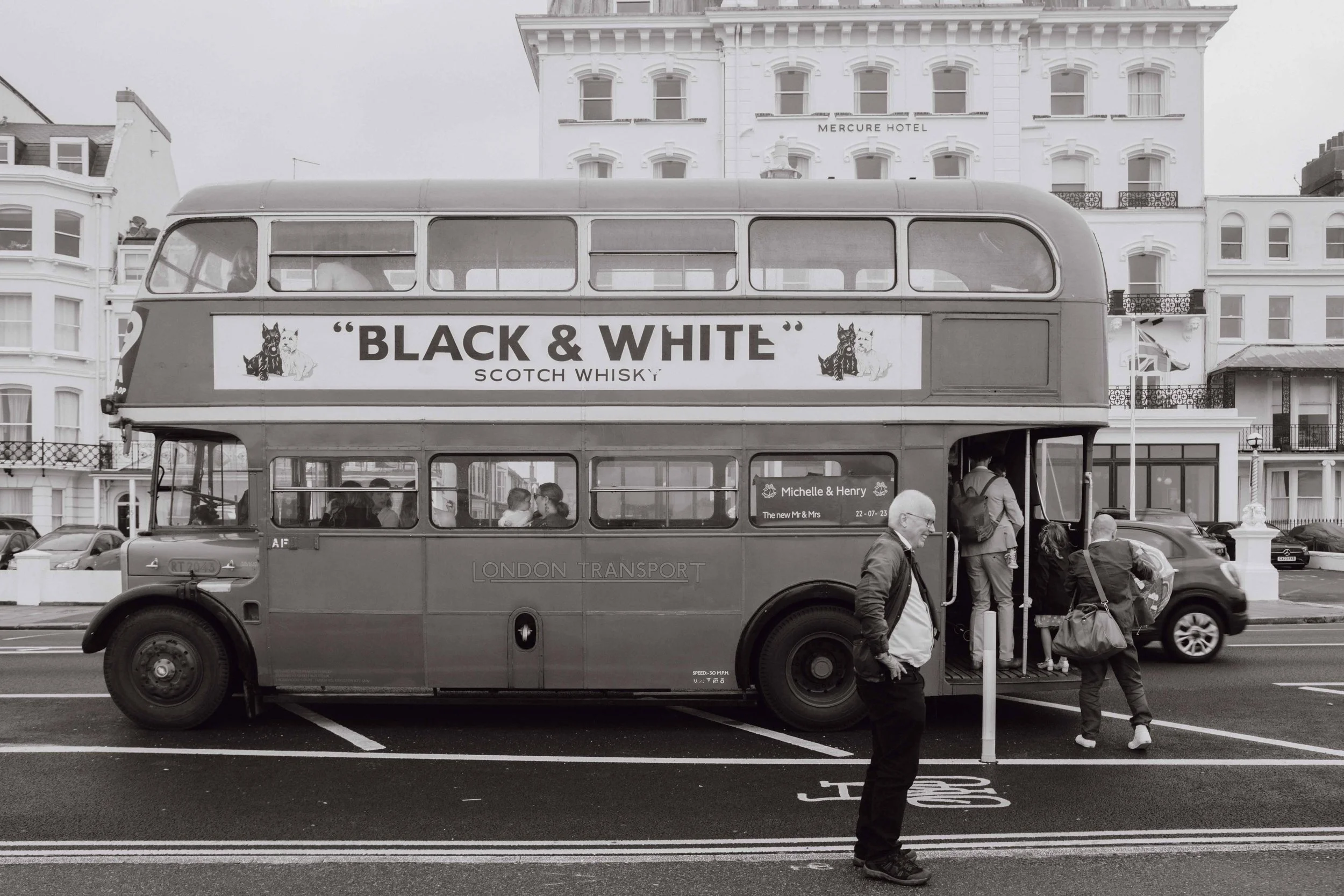 A vintage double-decker bus parked on a city street in front of white buildings. The bus has an advertisement for Scotch whisky called 'Black & White'. There are several people boarding and standing nearby, with a man in the foreground looking away f