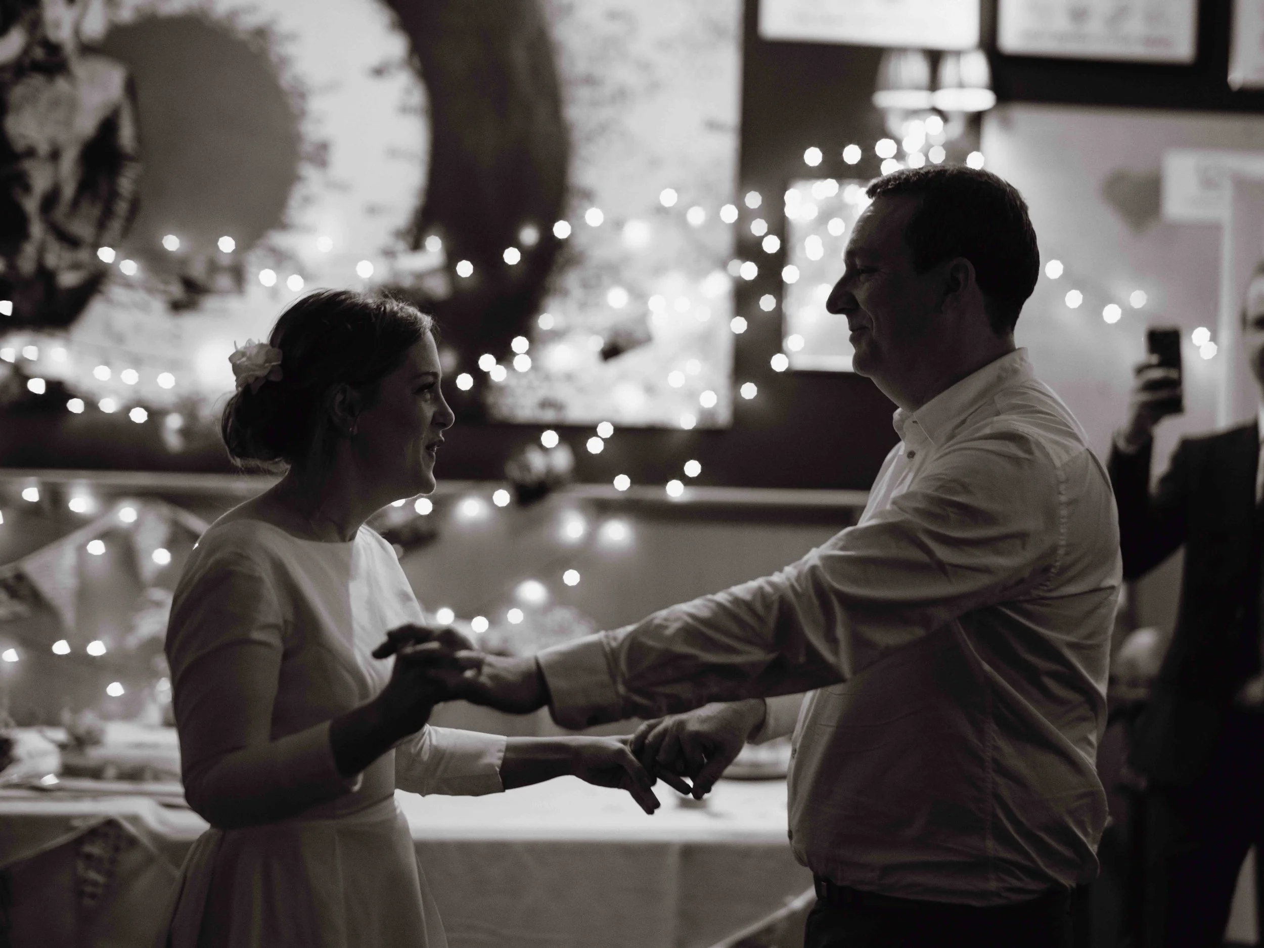 A couple dancing at a celebration, with string lights and artwork in the background, in black and white.