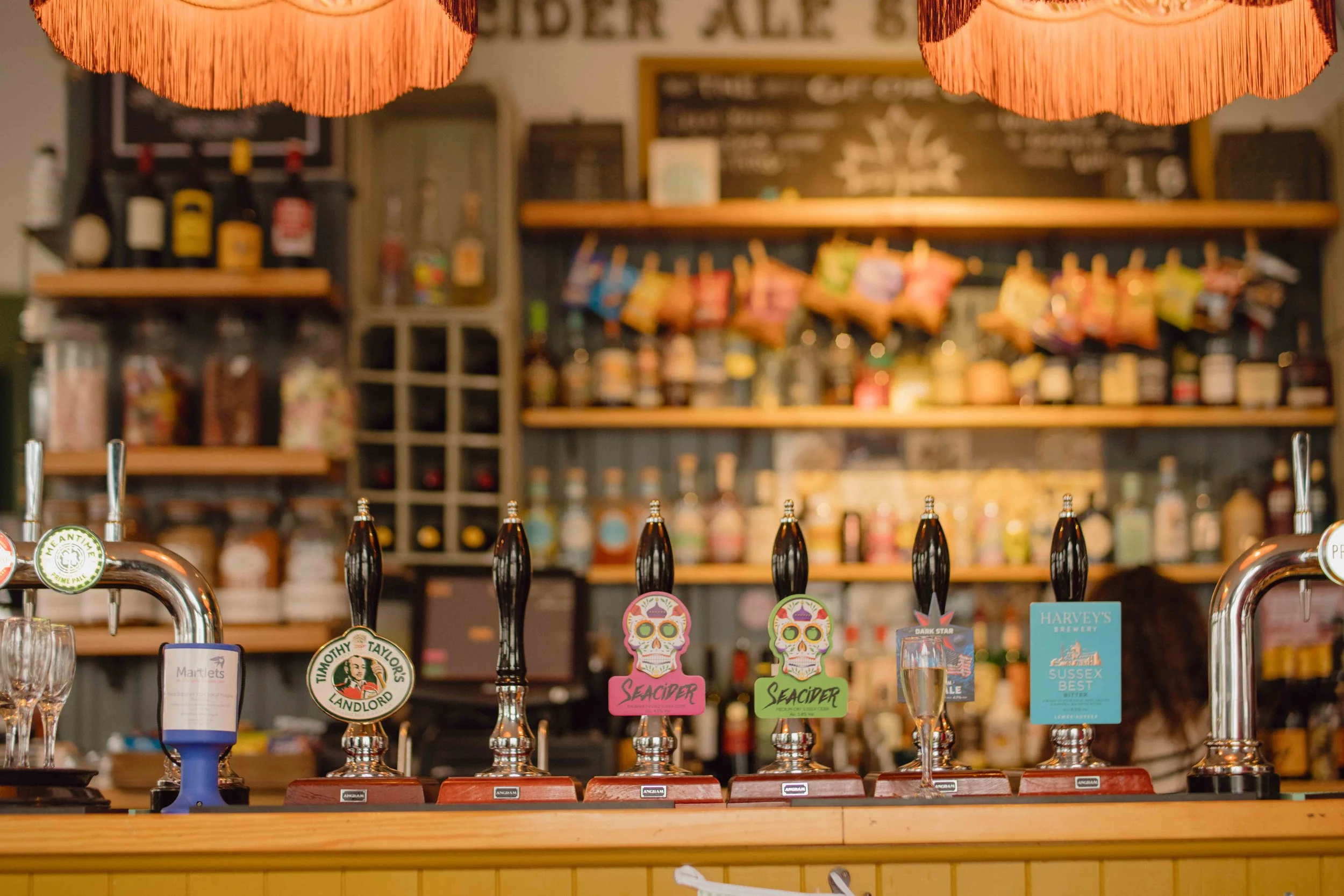 Bar with five beer taps featuring colorful skull labels, in a cozy pub setting with shelves of bottles and snacks in the background.