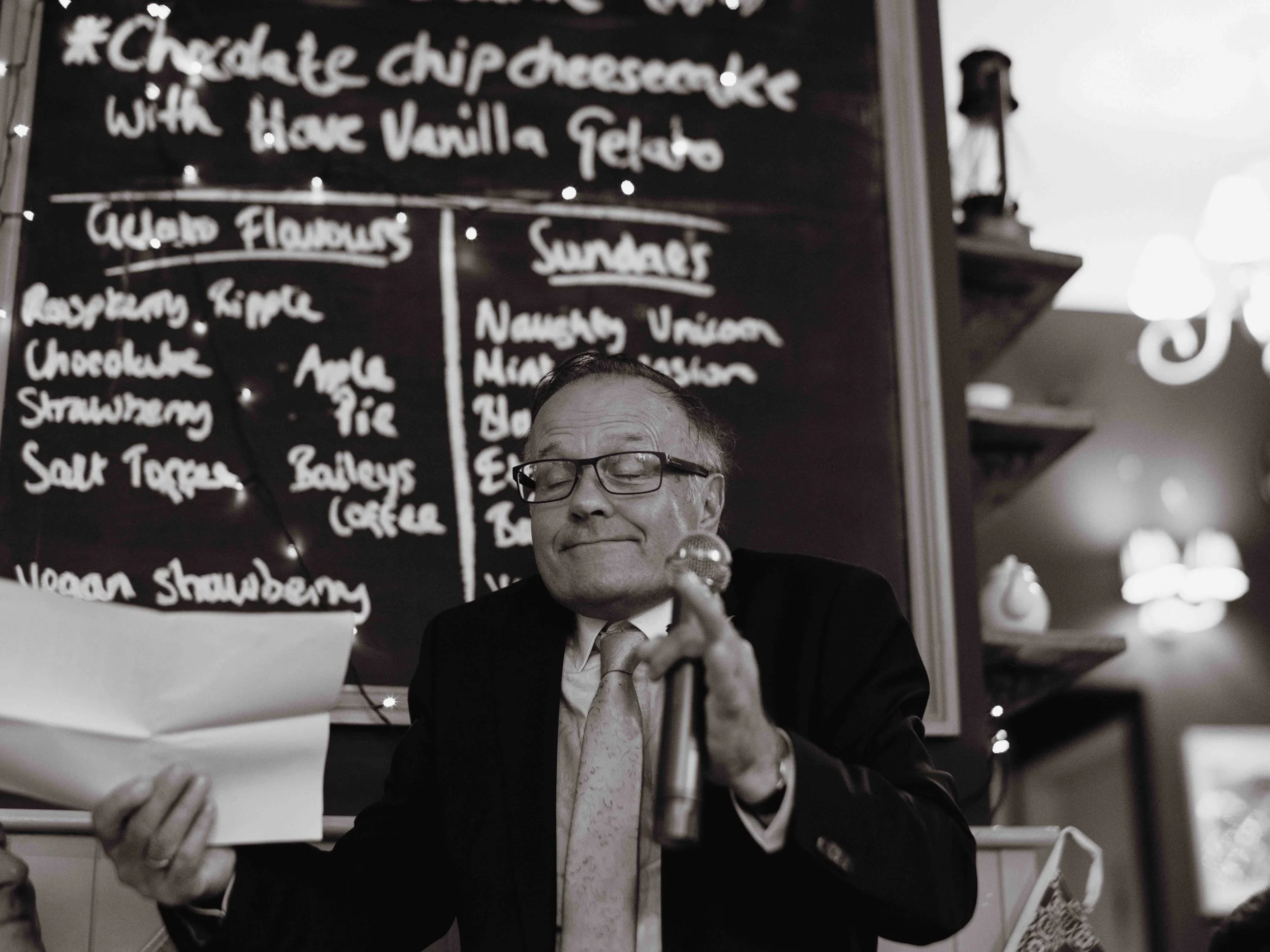 A man in a suit and glasses sitting at a table, holding a microphone, with a menu board in the background.