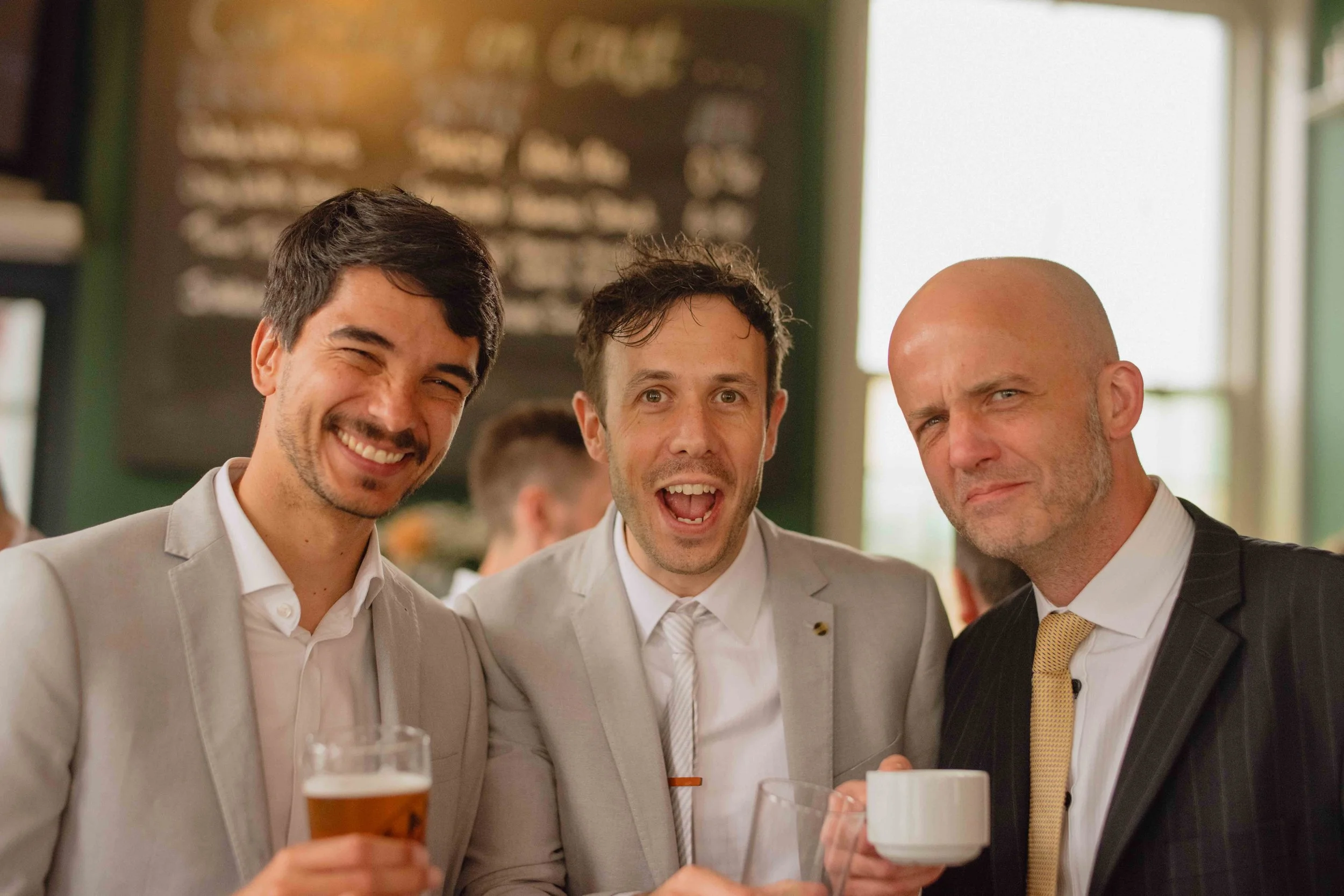 Three men in suits smiling and holding drinks at a social gathering in a restaurant or bar.