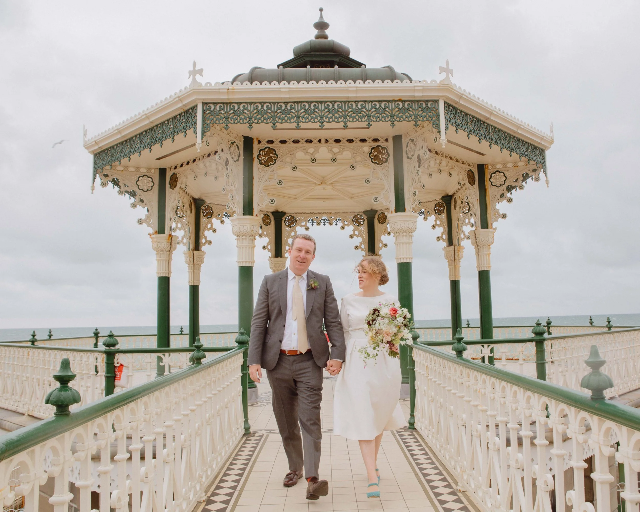 A newly married couple walking hand in hand under a decorative gazebo on a pier, with the sea and cloudy sky in the background.