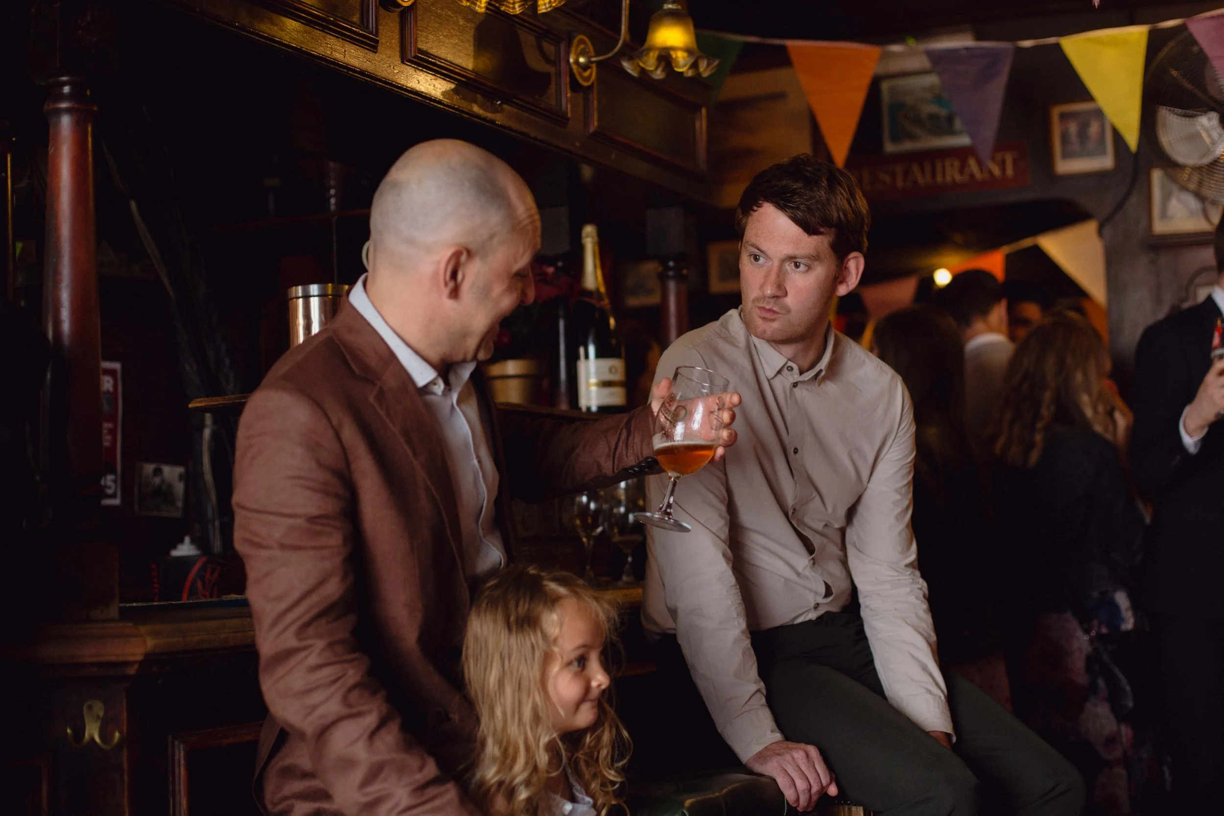 A man with a shaved head wearing a brown jacket holding a glass of drink, sitting next to a young girl with curly blonde hair, and another man with dark hair in a light-colored shirt sitting beside him in a crowded bar or pub with bunting and framed 
