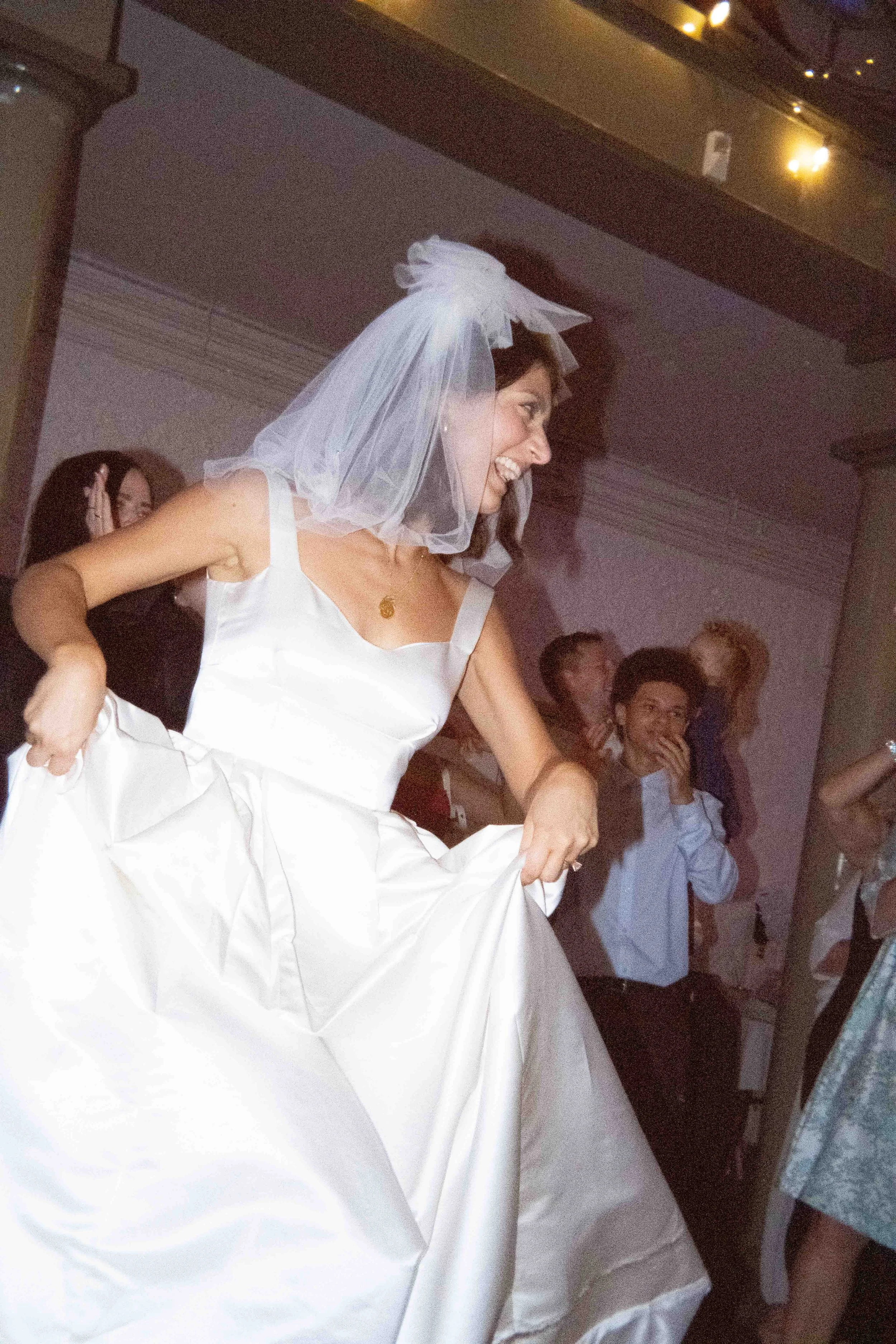 Bride dancing, wearing a white wedding dress and veil, smiling and holding her dress at a celebration with guests around.